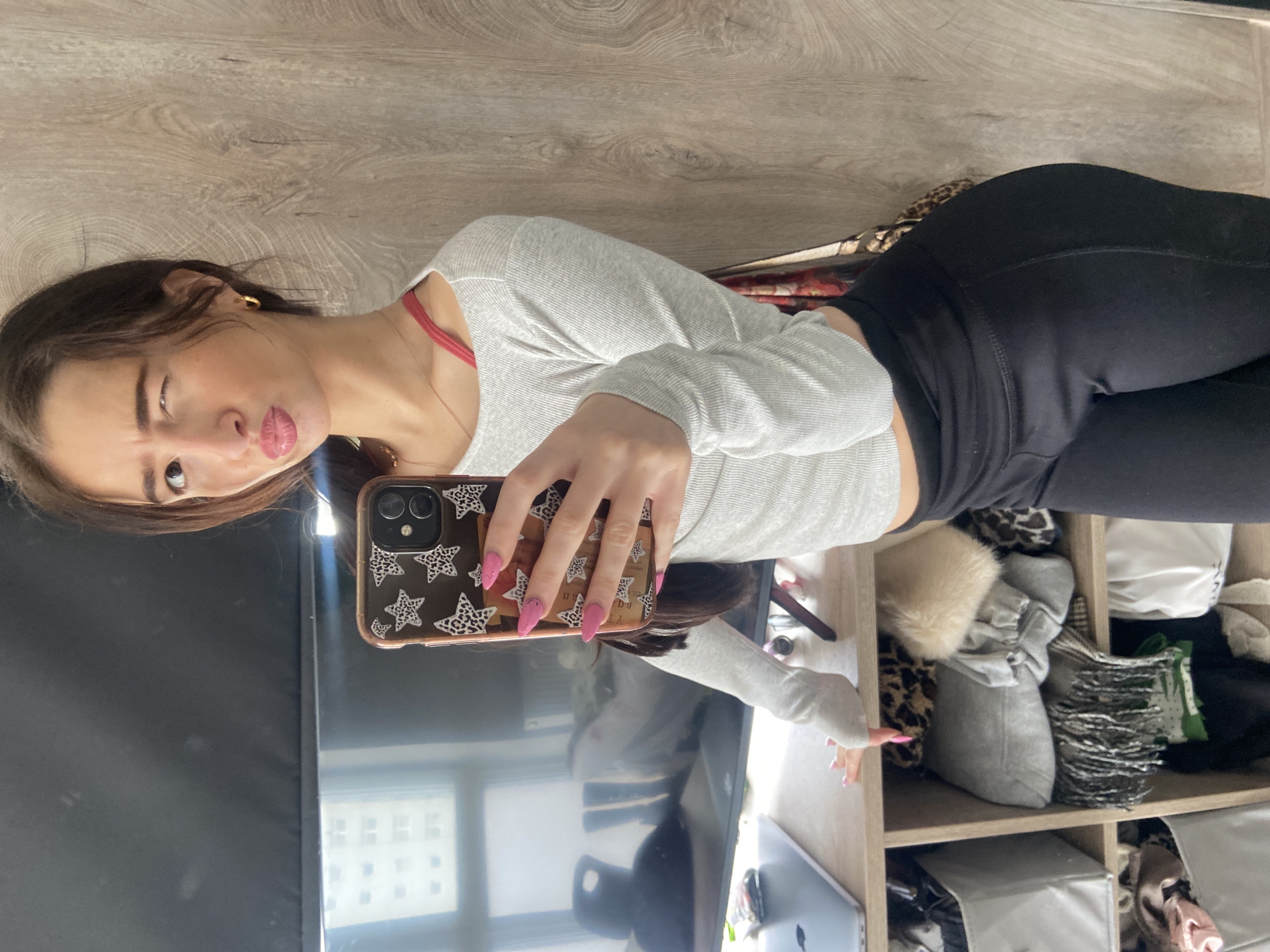 A woman is taking a selfie in a room with wood-paneled walls. She is winking at the camera and puckering her lips. She is wearing a light grey long-sleeved shirt, black leggings, and a red bra strap is visible under the shirt. She has pink manicured nails and is holding a phone with a clear case decorated with black and white star patterns. The phone reflects a black television screen and a window showing a building in the distance. A shelf unit filled with miscellaneous items, including a fur pillow, is visible to the right of the woman. Without additional specific details, I cannot confidently locate the location in London.