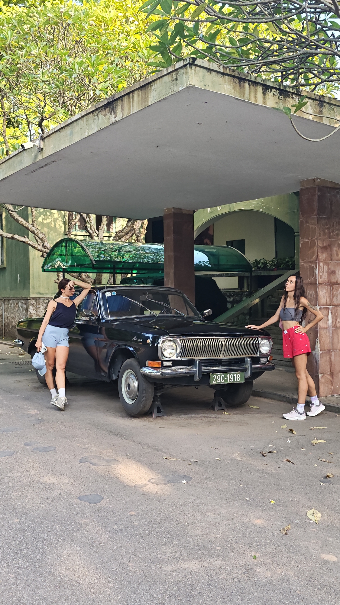 Two women are posing beside a black vintage car. The car is resting on jacks, suggesting it may be under repair. The location appears to be in Hà Nội, Vietnam. The license plate reads "29C-1918", which confirms the Hà Nội region. The women are casually dressed, and the setting seems to be a shaded area, possibly under a building's overhang. The environment includes trees and other architectural features, suggesting an urban setting.