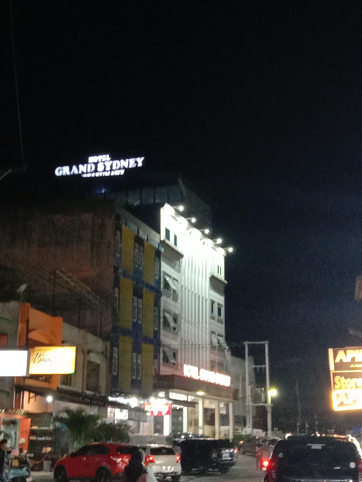 The image shows a nighttime street scene in Lhokseumawe, Indonesia, featuring the "Hotel Grand Sydney" prominently. The hotel building is visible, with its lighted sign on top. A mix of other commercial buildings and street lighting are apparent. Cars are on the road, indicating moderate traffic. People are present near the street and in front of the businesses.