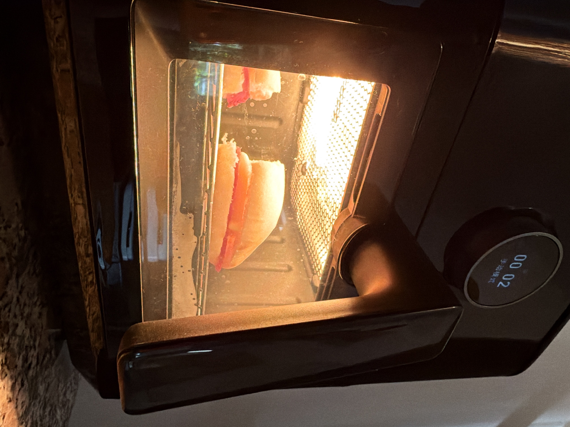 The image shows an open air fryer oven with two pieces of food being cooked inside on a rack. The food appears to be round and light colored, possibly buns for a sandwich, with a pink filling visible. The oven interior is lit by a bright heating element. On the exterior of the oven, a digital display shows "00 02" and Chinese characters that translate to "manual mode". The air fryer is black with a silver handle. The background is dark and out of focus, but appears to be a countertop. There are no people visible in the image. This scene is not specific to any location in the city of Ko Samui District, Thailand.