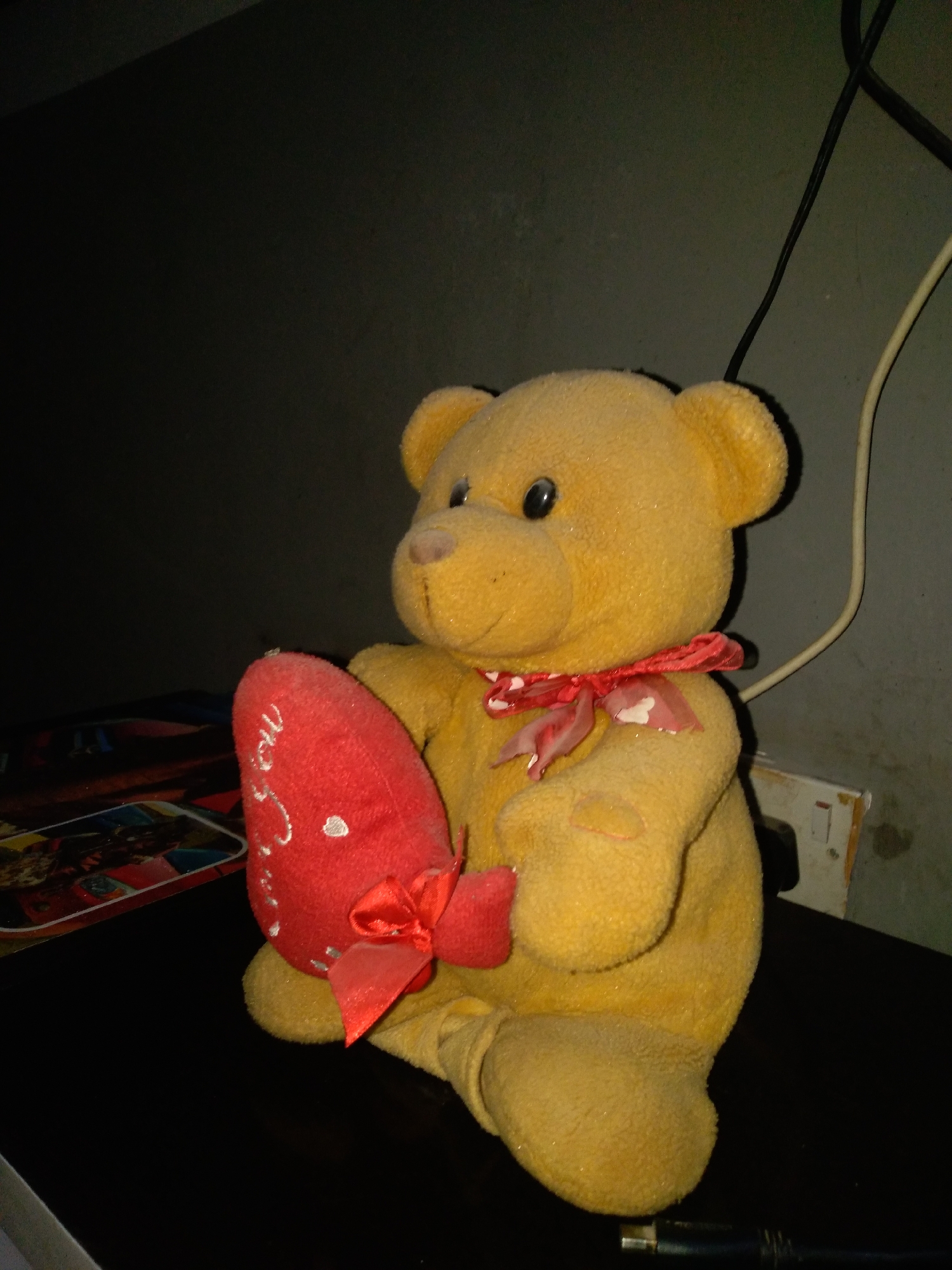 A close-up, low-angle shot shows a yellow stuffed teddy bear sitting on a dark surface, likely a table. The bear is holding a red heart-shaped pillow. The pillow has white embroidery that appears to be text and small white dots. The bear is wearing a red ribbon tied around its neck, with a bow and two dangling ends. The background is dark and out of focus, with some indistinct objects and wires visible. A portion of a white electrical outlet is visible on the right side of the frame. The lighting is dim, with some highlights on the bear's fur. The overall scene is indoors, in an unidentifiable indoor location.