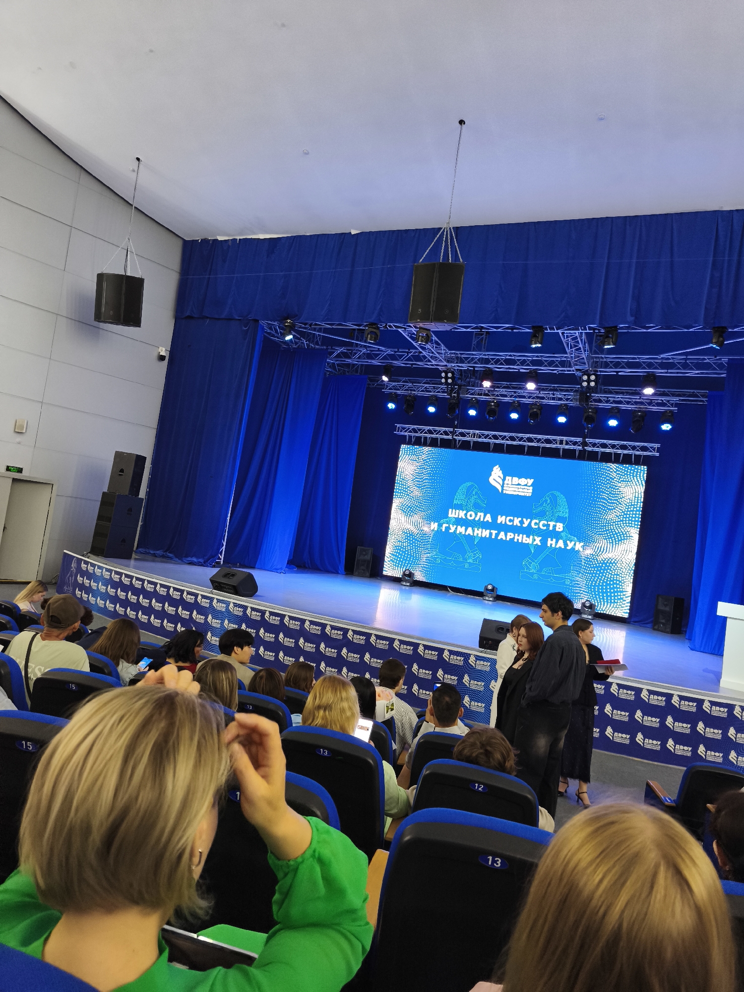 This image captures an indoor event, likely an awards ceremony or presentation, taking place in an auditorium. The stage is dominated by a large LED screen displaying a blue background with white text that reads "ШКОЛА ИСКУССТВ И ГУМАНИТАРНЫХ НАУК" (School of Arts and Humanities) and a logo above it. Blue curtains hang behind the screen, and stage lighting rigs are suspended above.
In front of the stage, a group of individuals is gathered, including several women and men. One woman in the foreground is wearing a bright green top. The audience is seated in rows of blue chairs, many of which are occupied by people of various ages. Some audience members are facing the stage, while others are looking at their phones or conversing.
The auditorium features a high ceiling with exposed structural elements and suspended speakers. Speakers are also visible on the left side of the stage, stacked vertically. The floor in front of the stage appears to be a dark color, and a white podium is situated on the right side. The backdrop for the audience seating area is a wall decorated with a repeating pattern of logos. The overall atmosphere suggests a formal or semi-formal gathering. The location is identified as Russky, Russia.