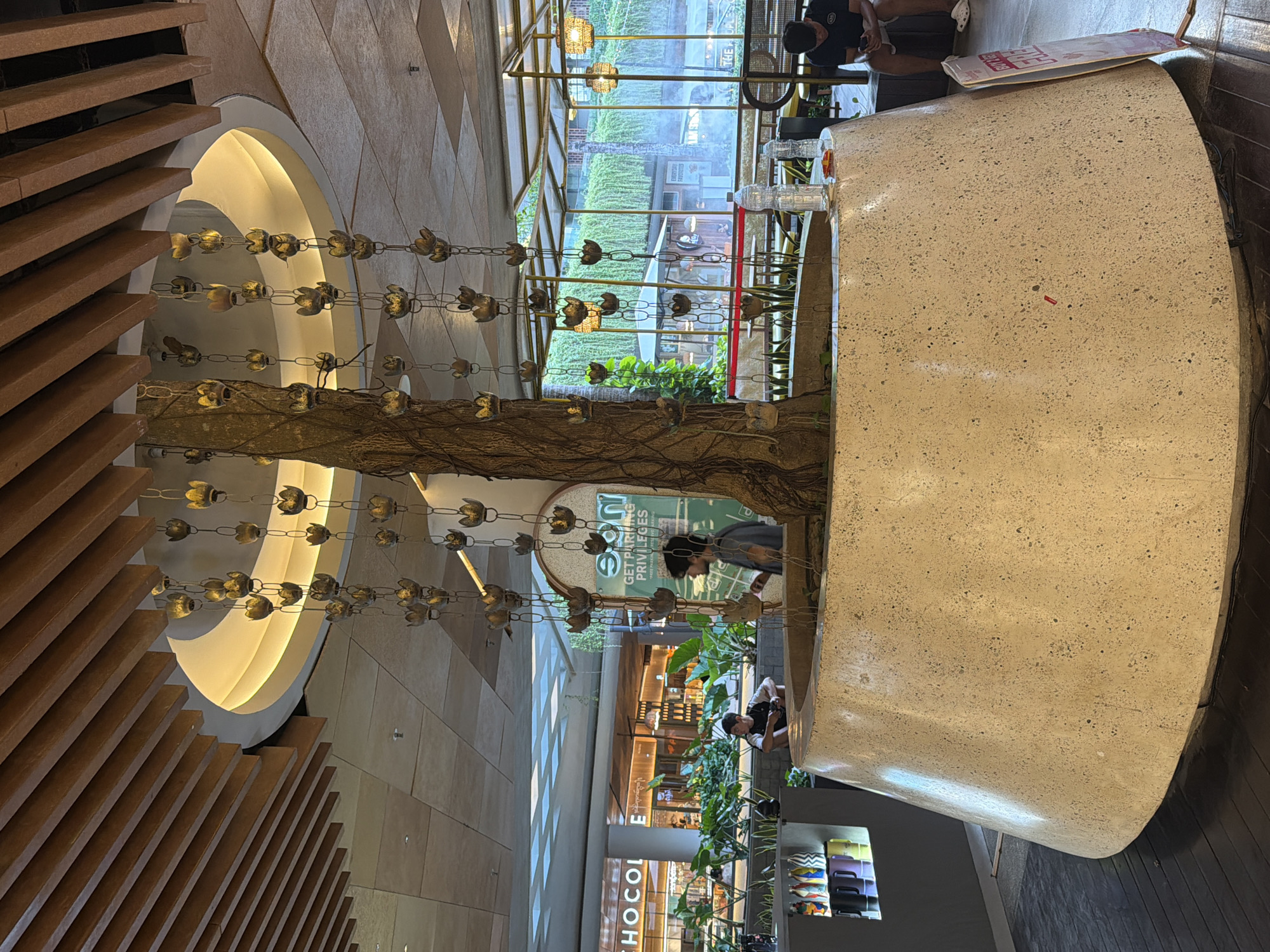 The image shows an interior space in Kuta, Indonesia, featuring a circular structure that acts as a centerpiece. A tree grows within this structure, its branches intertwined with chains of decorative metal flowers. Two people are positioned around the structure, one appearing to be using a cell phone. Beyond the circular structure, a view extends through glass windows or a storefront facade to an exterior area with greenery and other buildings, including one with the word "CHOCO." The interior design includes wooden slats on one side and patterned tile work on the other. Signage is visible but not fully legible.