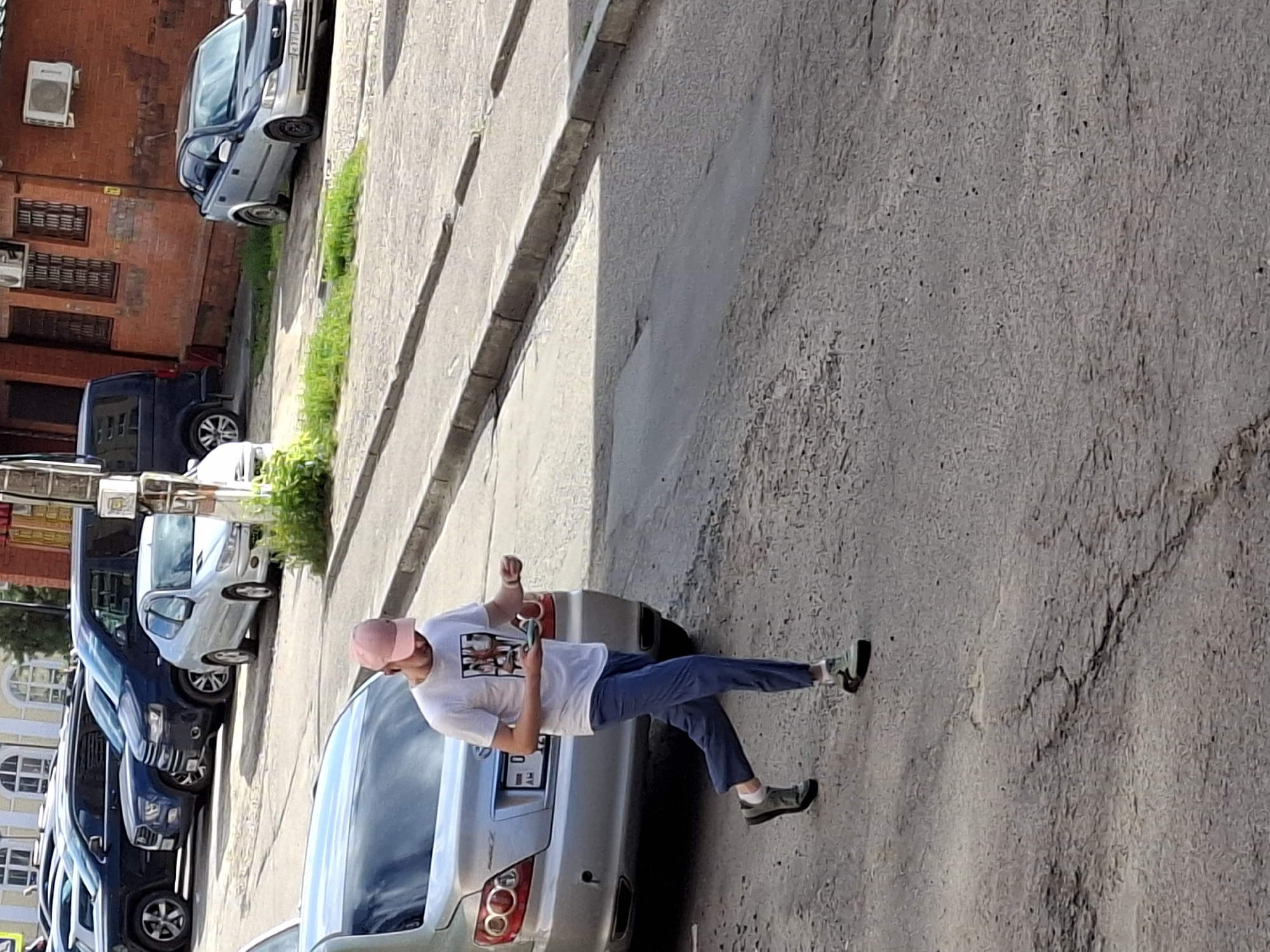 In an urban environment, a light-skinned male wearing a pink baseball cap, a white t-shirt with a graphic print, blue trousers, and dark-colored sneakers is walking on a cracked asphalt road. He is holding a dark-colored object, possibly a mobile phone, in his right hand and appears to be looking down at it. In the background, several parked cars are visible, including a silver sedan in the foreground to the left, and a row of SUVs and a minivan further down the street. A brick building with windows and an air conditioning unit is present on the right side of the image. The ground is uneven with patches of grass growing along the curb. The scene is illuminated by daylight.