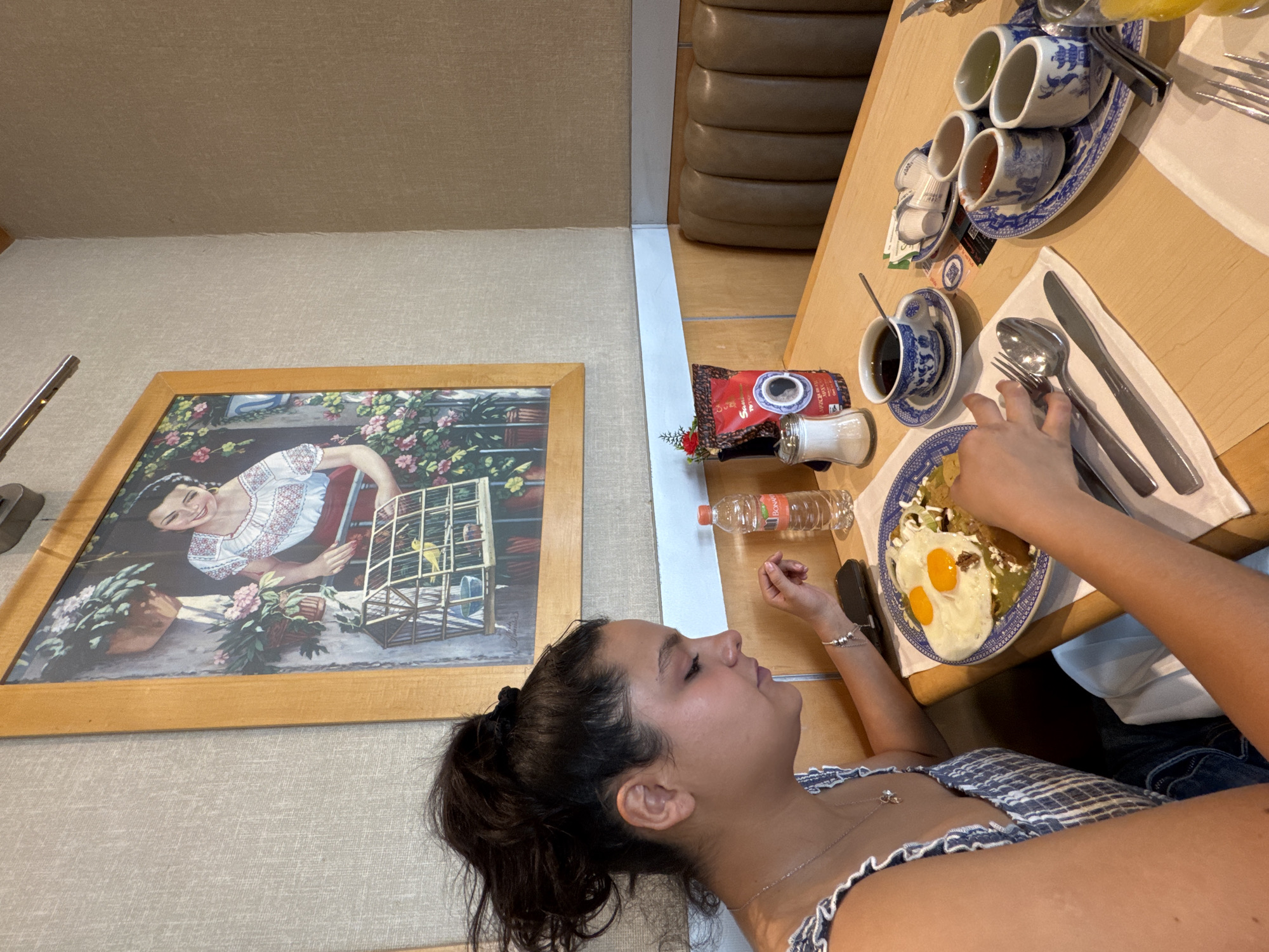 A woman is sitting at a table, about to eat a plate of chilaquiles with fried eggs. The table also has a set of blue and white China, a coffee cup and saucer, a bottle of water, a sugar packet, and a packet of coffee. A painting is on the wall, of a woman with a birdcage. The location is likely a restaurant in Puebla, Mexico, given the common local cuisine.