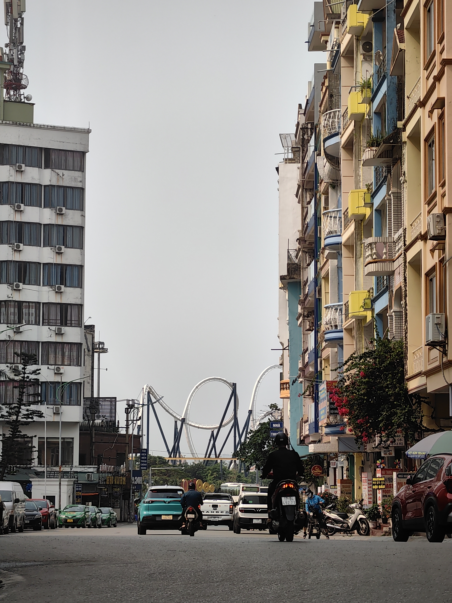 The image depicts a street scene in Bai Chay Ward, Vietnam. A roller coaster is visible in the distance between two rows of buildings. A variety of vehicles are traveling on the road, including cars, a motorcycle, and a truck. The buildings lining the street are a mix of colors and styles, some with balconies and air conditioning units. Street signs and shops are also visible along the street.