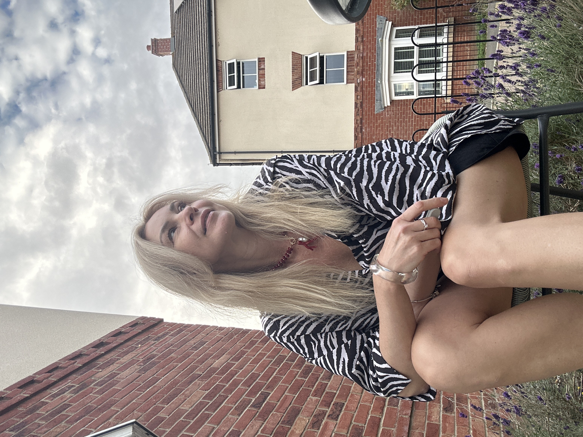 A medium shot shows a blonde woman with fair skin and visible makeup sitting outdoors. She is wearing a black and white zebra-print top with a low neckline, a red beaded necklace with a red coral pendant, and a silver bracelet. Her left hand rests on her knee, and her right hand is in her lap, wearing a silver ring. She is looking up and to her right, with her mouth slightly open.
The background consists of a red brick building on the left, a pale yellow house with brick accents on the right, and a cloudy sky above. The house has a dark roof, multiple windows, and a bay window on the ground floor. In front of the house, there is a black metal fence and a patch of lavender flowers. The woman is seated on a dark chair, partially visible. The overall scene appears to be in a residential area.