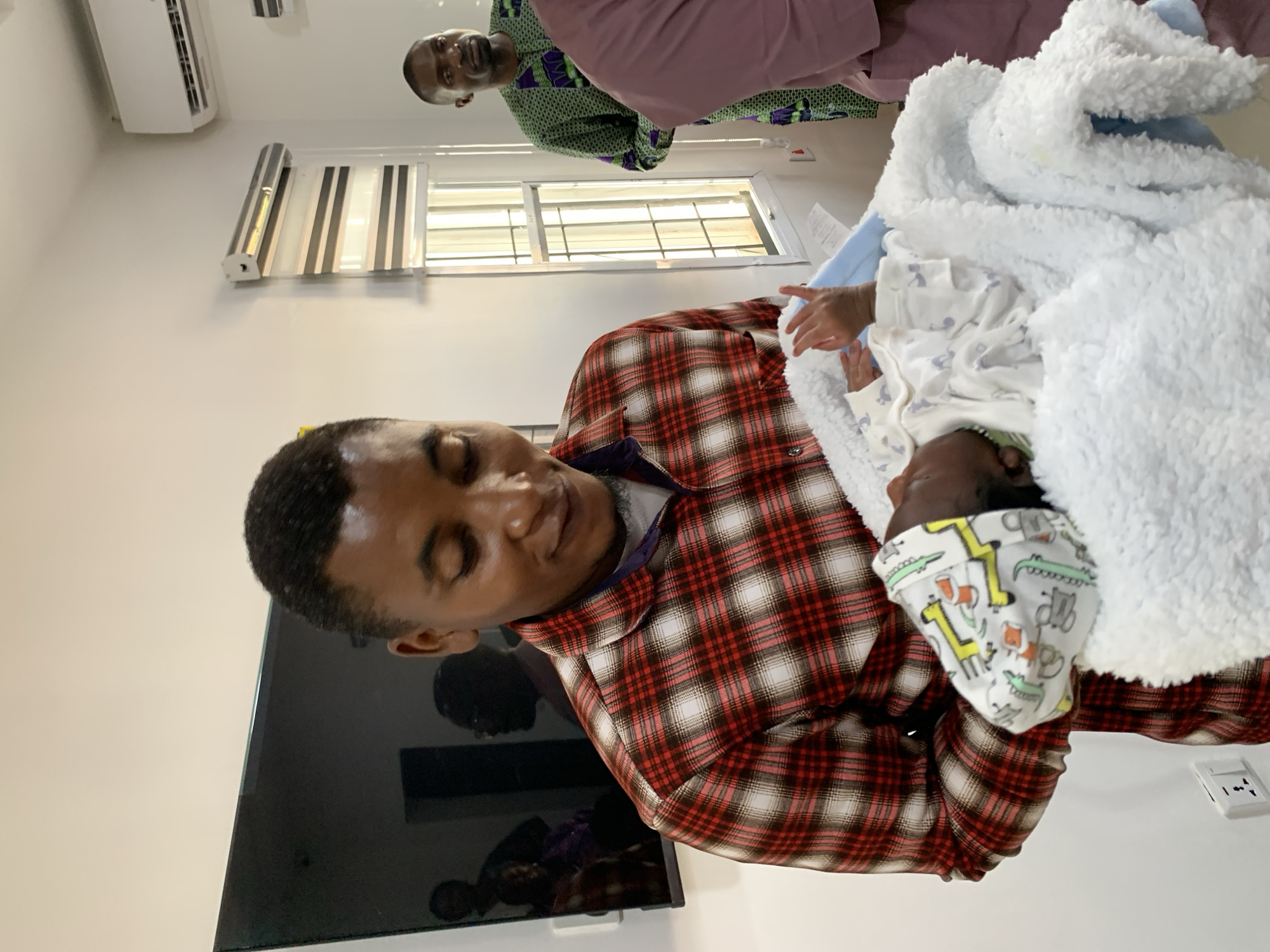 In an indoor setting, possibly a medical facility or a home in Jos, Nigeria, a Black male adult is holding a newborn baby. The adult, wearing a red, white, and brown plaid shirt over a purple undershirt, cradles the infant, who is wrapped in a white fluffy blanket with a light blue trim. The baby's left hand, with visible fingers, extends slightly from the blanket. In the background, another Black male adult wears a green and purple patterned garment. To the far right, a portion of a person in a pink garment is visible. The environment includes a large, dark television screen mounted on the wall to the left, reflecting several figures, and a window with blinds behind the individuals. An air conditioning unit is mounted high on the wall. A white electrical outlet is present on the wall near the bottom left of the frame. The lighting appears to be artificial, creating a slightly warm ambiance.