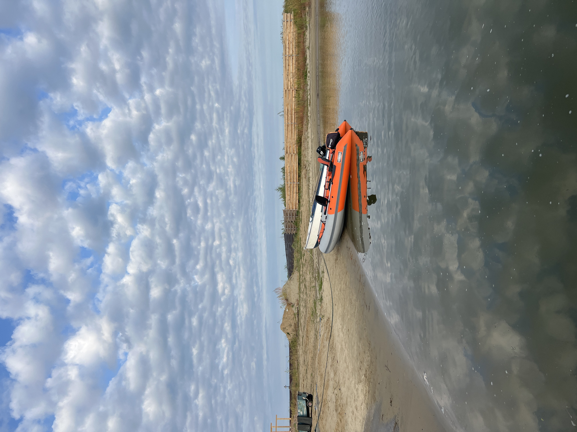 The image depicts a calm body of water bordering a sandy shore. Two inflatable boats are situated on the shore, partially submerged in the water. The foreground is dominated by the tranquil water, reflecting the cloudy sky above. The sandy shore features scattered debris, including a green trailer with a black tarp, and a dark rope extending across the sand and attaching to the boats. Further inland, a dirt mound is visible, bordered by a wooden fence and sparse vegetation. The sky is filled with a complex pattern of grey and white clouds against a blue backdrop. The scene appears to be a quiet, possibly undeveloped waterfront area. There are no people visible in the image. The location is identified as Ново-Пичуго, Russia.