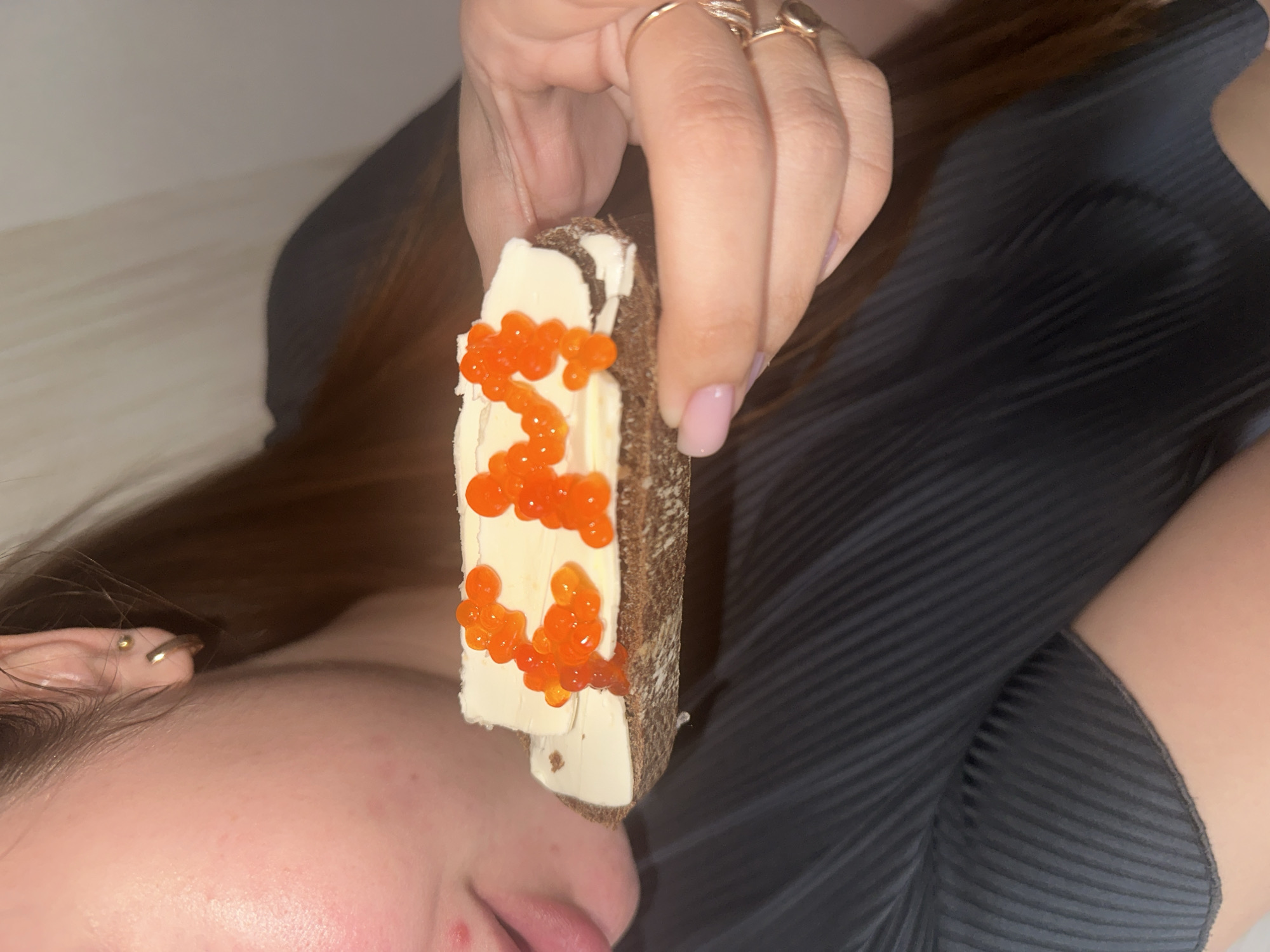 A woman lies on her side, close-up, holding a slice of dark bread covered with butter and salmon roe. The location cannot be determined from the image.