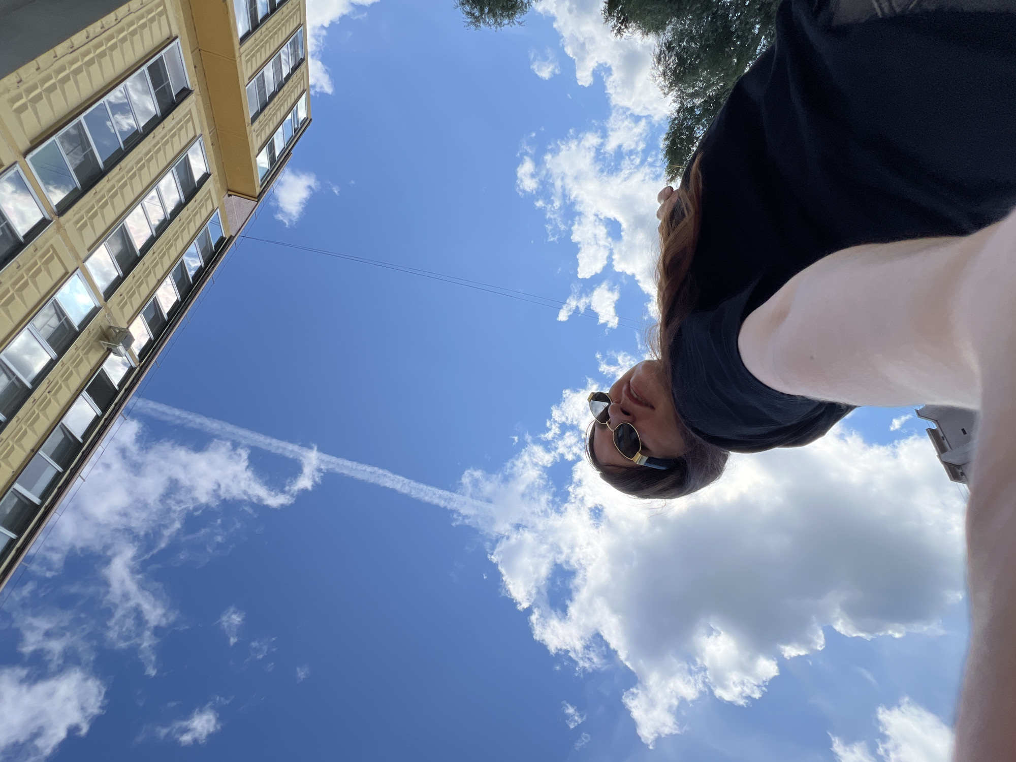 The image is a low-angle, upside-down selfie taken outdoors in Noviy, Russia, on a clear day with fluffy white clouds. A young woman with long brown hair and sunglasses is the main subject, looking upwards. She is wearing a black t-shirt and appears to be standing near a building. The building, partially visible at the top of the image, is a multi-story structure with numerous windows. The overall scene suggests a bright, daytime setting.