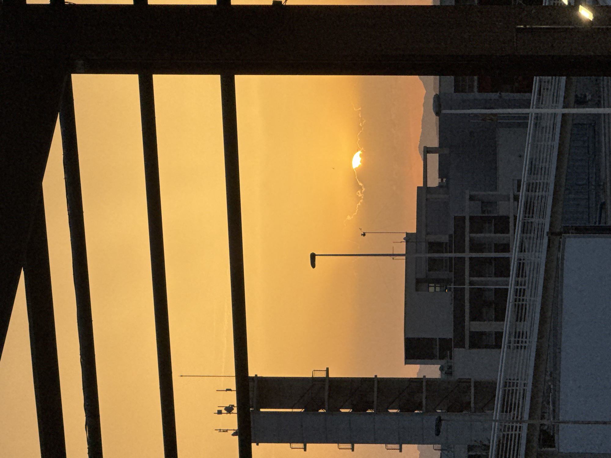 The image depicts a view through dark horizontal bars against an orange sky, characteristic of a sunset in Torrevieja, Spain. The scene is framed by these bars, possibly part of a building's facade or structure, suggesting an urban setting. In the distance, the sun is partially obscured by clouds, casting a warm glow. Silhouetted against the sky are several buildings, including a tall, rectangular structure with balconies, likely a tower, and other multi-story buildings. Antennae and streetlights are also visible. A pedestrian bridge with white railings spans across the lower part of the frame. The overall impression is an urban landscape at dusk, with the natural beauty of the sunset juxtaposed against the man-made structures.