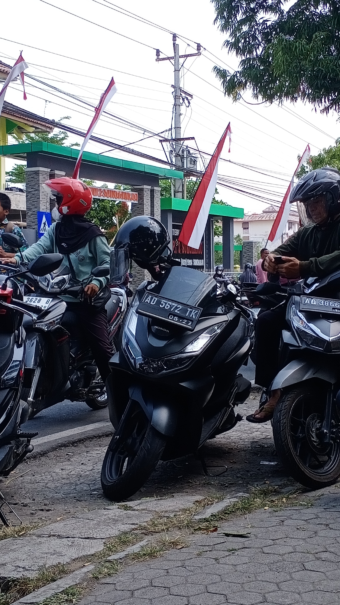 In Joho, Indonesia, a medium shot captures a street scene with several motorcycles and a few individuals. The foreground is dominated by two black scooters. The scooter on the left displays a license plate that reads "3628 B". The scooter on the right has a license plate "AD 5672 TK" and the word "HONDA" is visible below the plate. A person wearing a dark green jacket and a black helmet is seated on this scooter, actively using a mobile phone. The scooter directly to the right of this one has a license plate that reads "AD 3866".
Behind these scooters, a person wearing a red helmet and a light blue jacket with black headscarf is seated on another motorcycle. This individual appears to be looking forward, and a black bag is slung across their chest. Several Indonesian flags, red and white, are prominently displayed on poles, some attached to a structure that appears to be an entrance or archway. This archway has text visible on it, including "SUKOHARJO". Overhead, power lines and utility poles are visible against a bright sky, with some trees providing a backdrop. The ground is paved with interlocking stones. The overall atmosphere suggests a moment of pause, possibly traffic or a gathering, in a semi-urban environment decorated for a local event.