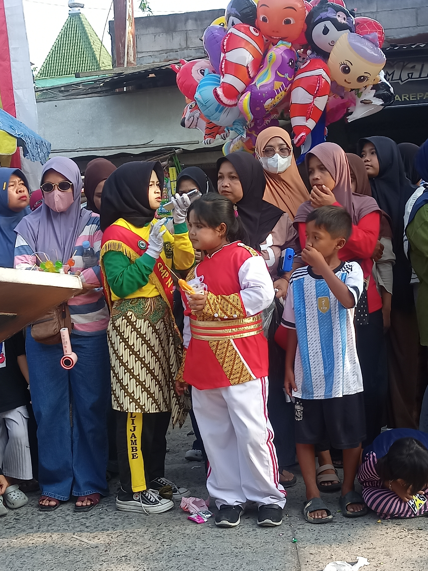 In the city of Saren, Indonesia, a group of children and adults are gathered outdoors. The scene appears to be a street or market area, with buildings and possibly a mosque in the background. Many of the people are wearing traditional Indonesian clothing, including hijabs.
In the foreground, a young girl in a red and white marching band uniform holds a cup and appears to be chewing on something. Beside her, a boy in an Argentinian soccer jersey stands with his hand to his mouth. Behind them, a group of women and children watch the festivities. A table with drinks and snacks is visible on the left side of the image. Several balloons, including some shaped like cartoon characters, are floating in the background. The overall atmosphere is festive and communal, suggesting a local celebration or event.