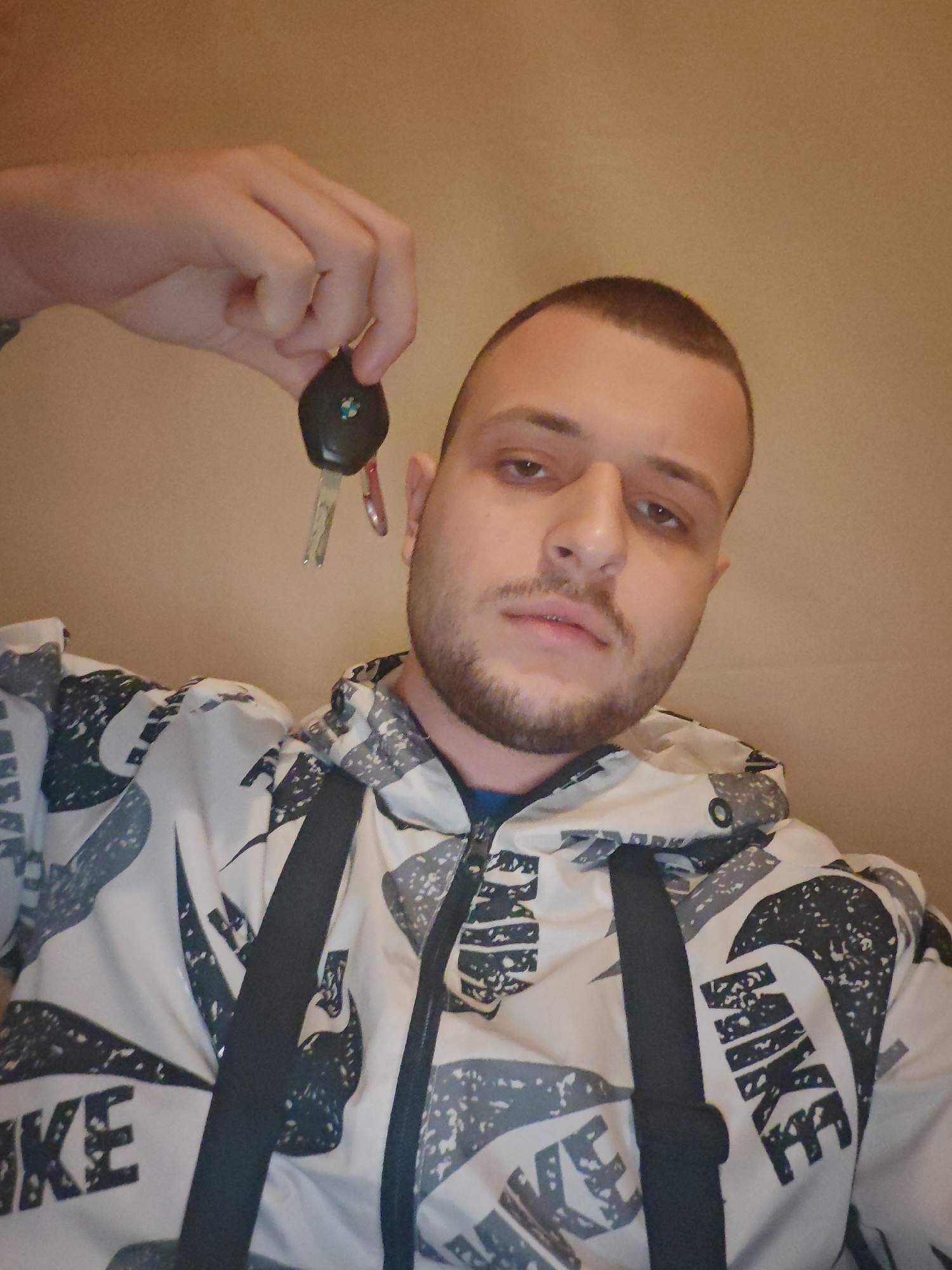A young man with short hair and stubble is holding up car keys in his right hand, which is extended upwards. He is wearing a white jacket with a black and grey camouflage pattern and large black "NIKE" logos. The background is a plain, light brown wall. The lighting suggests an indoor setting. No specific location in Burgas, Bulgaria can be determined from the image.