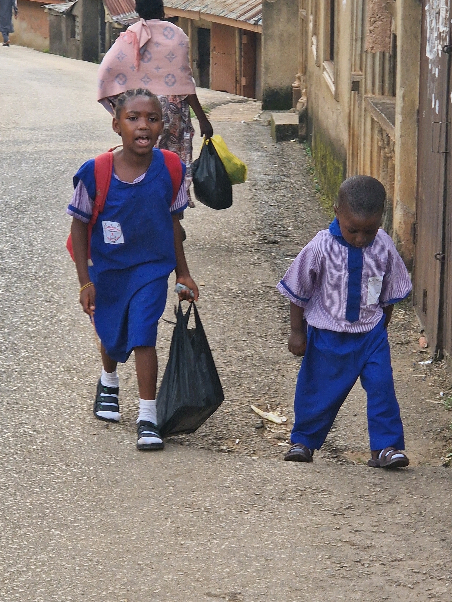 In the city of Jos, Nigeria, a young girl in a blue school uniform, with a red backpack, walks down a paved street. She holds a black plastic bag in her right hand. To her right, a younger boy in a striped and blue uniform walks, looking down at the ground. In the background, a woman in a pink headscarf and patterned dress walks away from the camera, carrying a yellow plastic bag. Buildings with corrugated metal roofs line the street. The scene appears to be during daylight hours, with overcast skies.