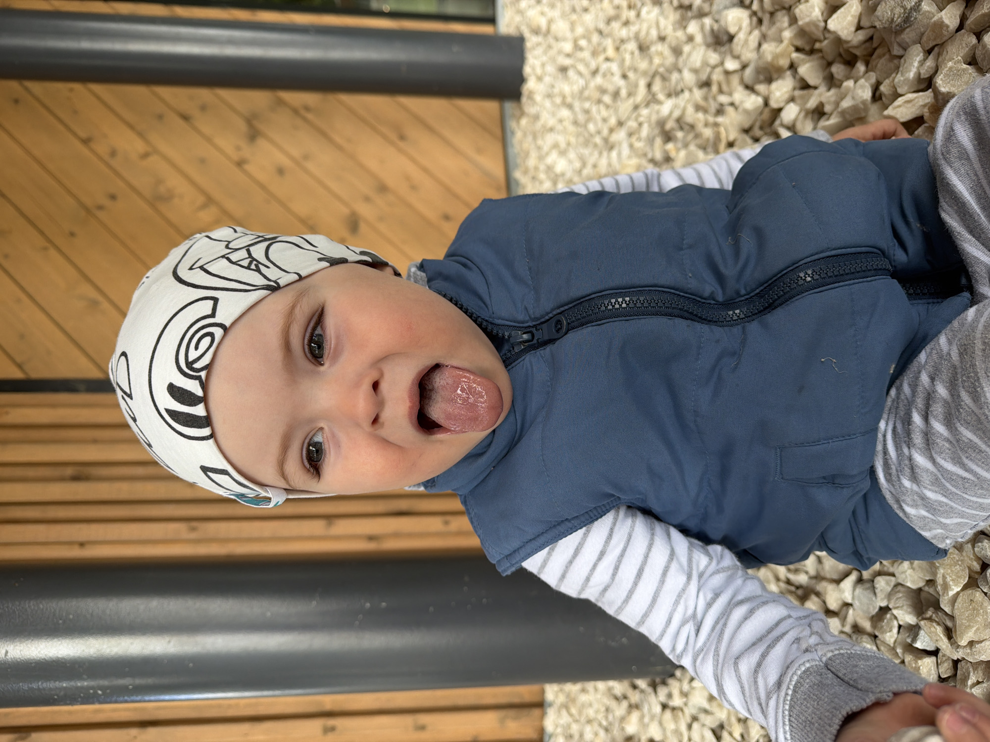 The image shows a close-up of a young child wearing a white beanie with black line drawings, a blue vest, and a striped long-sleeved shirt. The child is sticking their tongue out, with their mouth open. They are sitting on a surface covered with light-colored gravel. In the background, there is a wooden wall with vertical planks and a dark, cylindrical pole. The overall setting appears to be outdoors, possibly a park or a patio area. There are no discernible landmarks or features that would specifically place this image in Votkinsk, Russia.