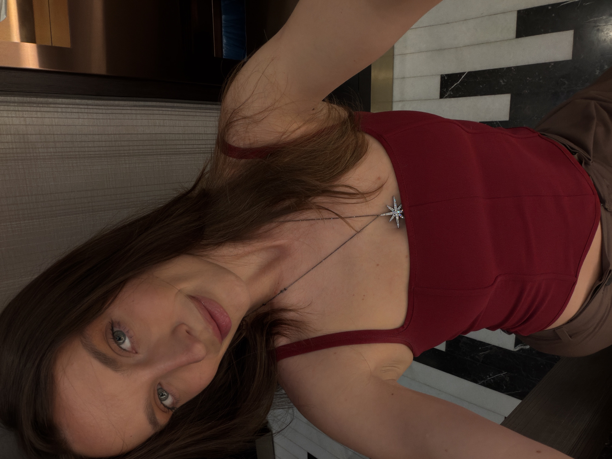 A woman with light skin and brown hair is centered in the frame, wearing a red tank top and brown pants. She also has on a silver necklace with a star-shaped pendant. The background reveals a room with a black and white tiled floor, likely indoors. The city of Budva, Montenegro, cannot be determined from this image.