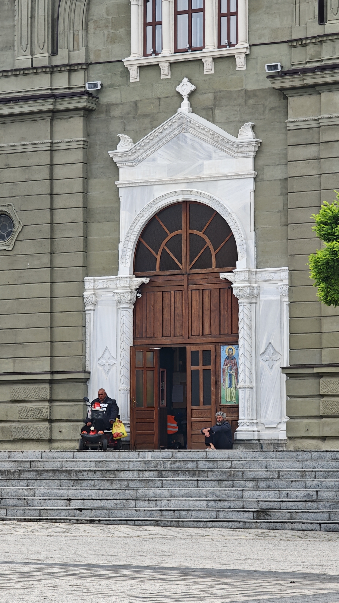 The image shows the entrance of the Saint Cyril and Methodius Cathedral in Burgas, Bulgaria. A man sits on a motorized scooter to the left of the entrance, holding a yellow shopping bag. Another man sits cross-legged on the steps to the right of the entrance. The cathedral doors are partially open, revealing an orange vest inside. A religious image is displayed on the door. The architecture features light gray stone with white marble accents around the doorway. A gray stone staircase leads up to the entrance. The foreground is a light-colored paved area.