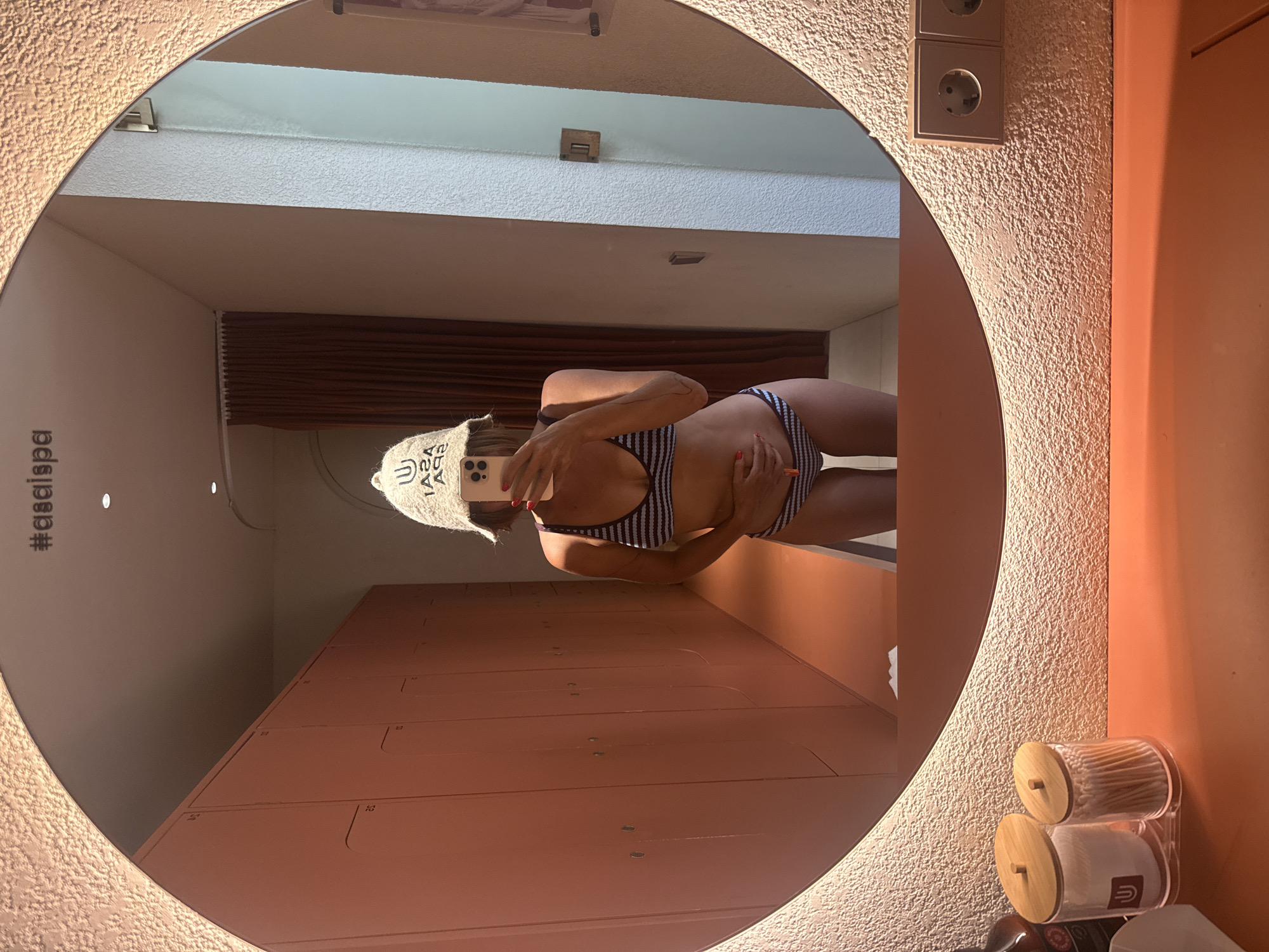 A person takes a mirror selfie in a brightly lit room. The mirror is round and framed with a textured, tan-colored material, and the room's walls are the same color. A double electrical outlet is visible on the wall near the mirror. The person is wearing a striped swimsuit and a white beanie. Behind the person, there are pink lockers lining the wall. The reflection shows the room's interior, including a white ceiling, brown curtains, and what appears to be a second wall behind the lockers. The lower right corner of the mirror shows a container with wooden lids. The location is Kuta Selatan, Indonesia.