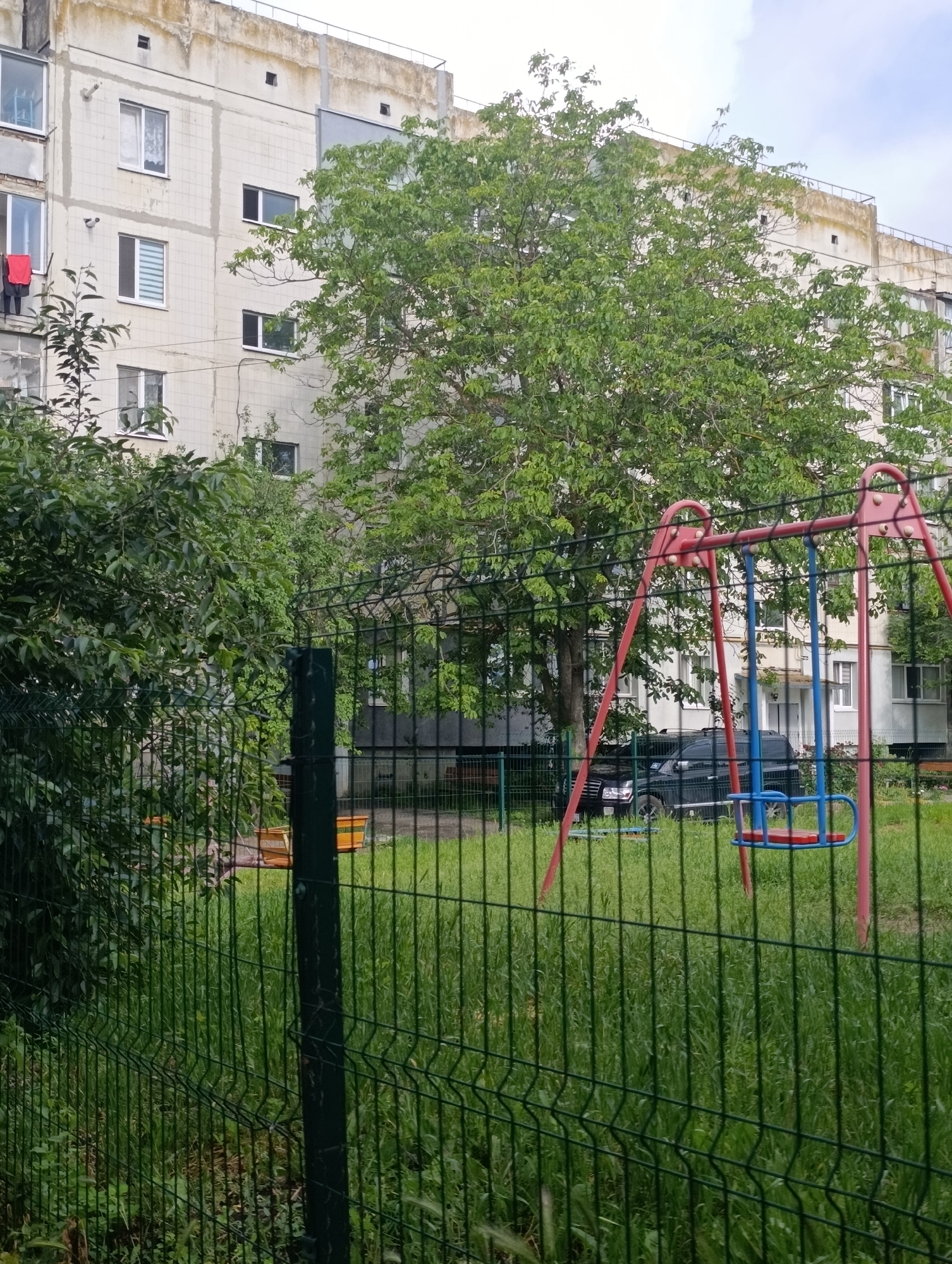 In Khorol, Ukraine, a multi-story apartment building with a rough, concrete facade stands behind a green wire fence. A large tree with dense green foliage is situated in front of the building, partially obscuring the windows. A red and blue swing set is visible in the foreground, positioned on a grassy area. A dark-colored SUV is parked behind the fence near the apartment building. Red clothing hangs from a line outside one of the windows on the left side of the building. The sky above is overcast.