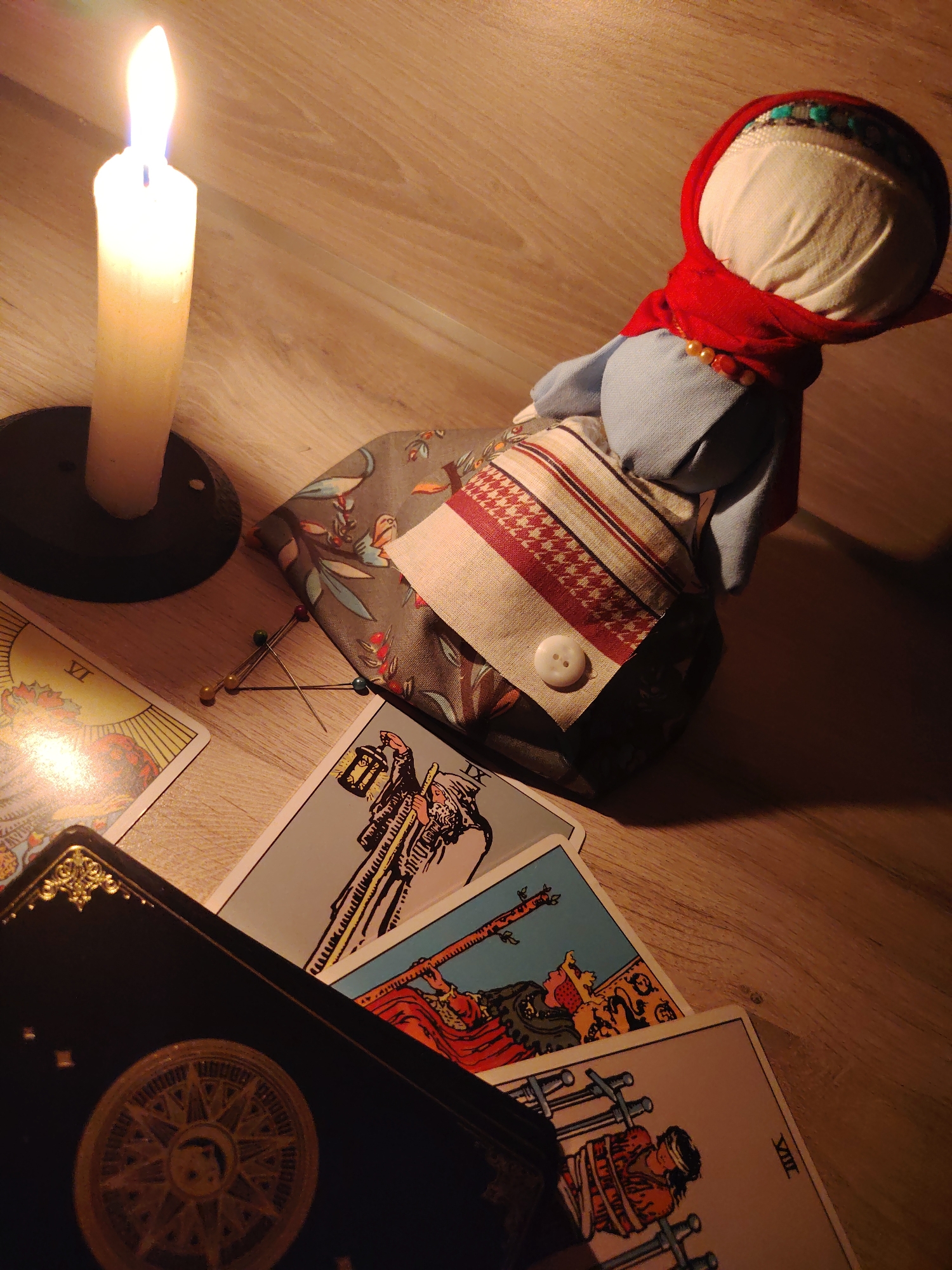 The image shows a dimly lit wooden surface with various objects related to divination or witchcraft. A tall, white candle burns in a black holder on the left side of the image. A cloth doll with a white head wrapped in a red scarf sits to the right of the candle. The doll is wearing a blue garment and a vest with a patterned fabric and a single white button. Several tarot cards are scattered around the doll and at the bottom of the frame. A black tarot deck box is partially visible in the lower-left corner, adorned with a golden compass or astrological symbol. A few pins are scattered near the doll. The lighting is warm and low, casting shadows across the scene. The location is unknown; there is no information provided within the image to indicate that it is in the city of Chaikovsky, Russia.