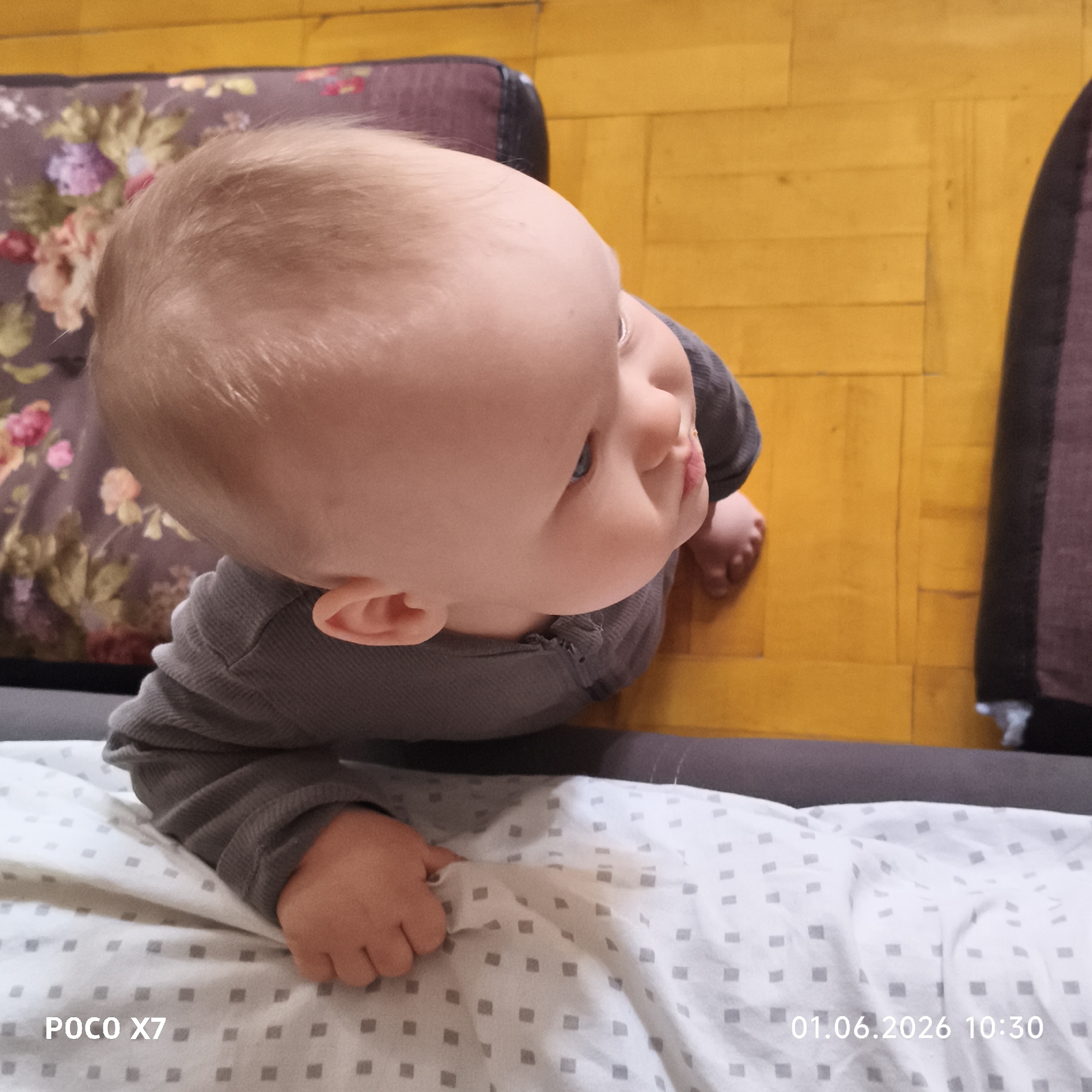 A high-angle shot shows a baby lying on a white, patterned blanket. The baby wears a gray long-sleeved shirt and is looking upwards and to the right. The baby's right hand is resting on the blanket. To the baby's left is a floral-patterned pillow, and on the right, a dark-colored pillow. The floor is made of yellow wooden planks. A "POCO X7" watermark is in the bottom left corner, and a date and time stamp "01.06.2026 10:30" are in the bottom right corner. The location is unknown.