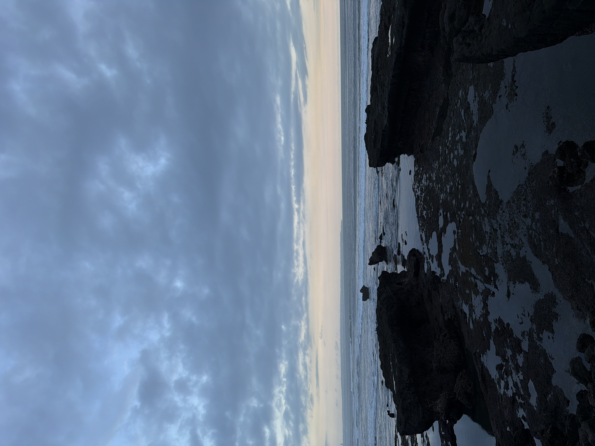 The photograph depicts a coastal scene, likely taken at dusk or dawn. The sky, covering the left side of the image, is filled with dark, layered clouds, suggesting an overcast day. Towards the horizon, a narrow strip of lighter, yellowish sky is visible. The ocean extends from the horizon to the center of the image, showing gentle waves meeting the shore.
On the right side, dark, rocky formations are visible, partially covered with water. The wet rocks reflect the sky, creating a mirrored effect. The scene appears tranquil, with no people or apparent activity.
Given the location of North Kuta, Indonesia, it's plausible this image shows a rocky outcrop along the coastline, likely a tidal area or a beach with volcanic rock formations, like those found in areas such as Canggu or Tanah Lot. Without a specific landmark or distinguishing feature, it is hard to provide a more exact location.