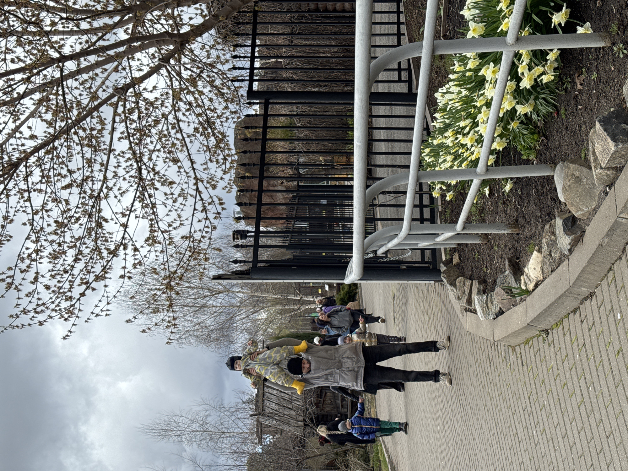 The image depicts a group of people on a brick walkway, near a metal fence and railing. A man is carrying a child on his shoulders. There are trees with budding leaves visible, as well as daffodils planted along the walkway. The photograph was taken on a cloudy day. The location is Izhevsk, Russia.
