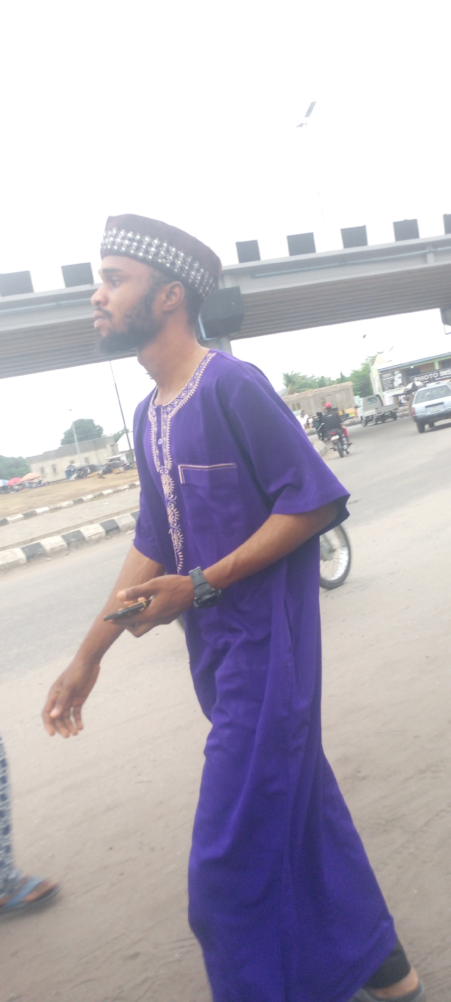 A man with a beard wearing a purple robe and a hat is walking on a street in Adankolo, Nigeria. He is holding a phone and wearing a watch. There is a road, bridge, and buildings in the background. There are other people and vehicles present.