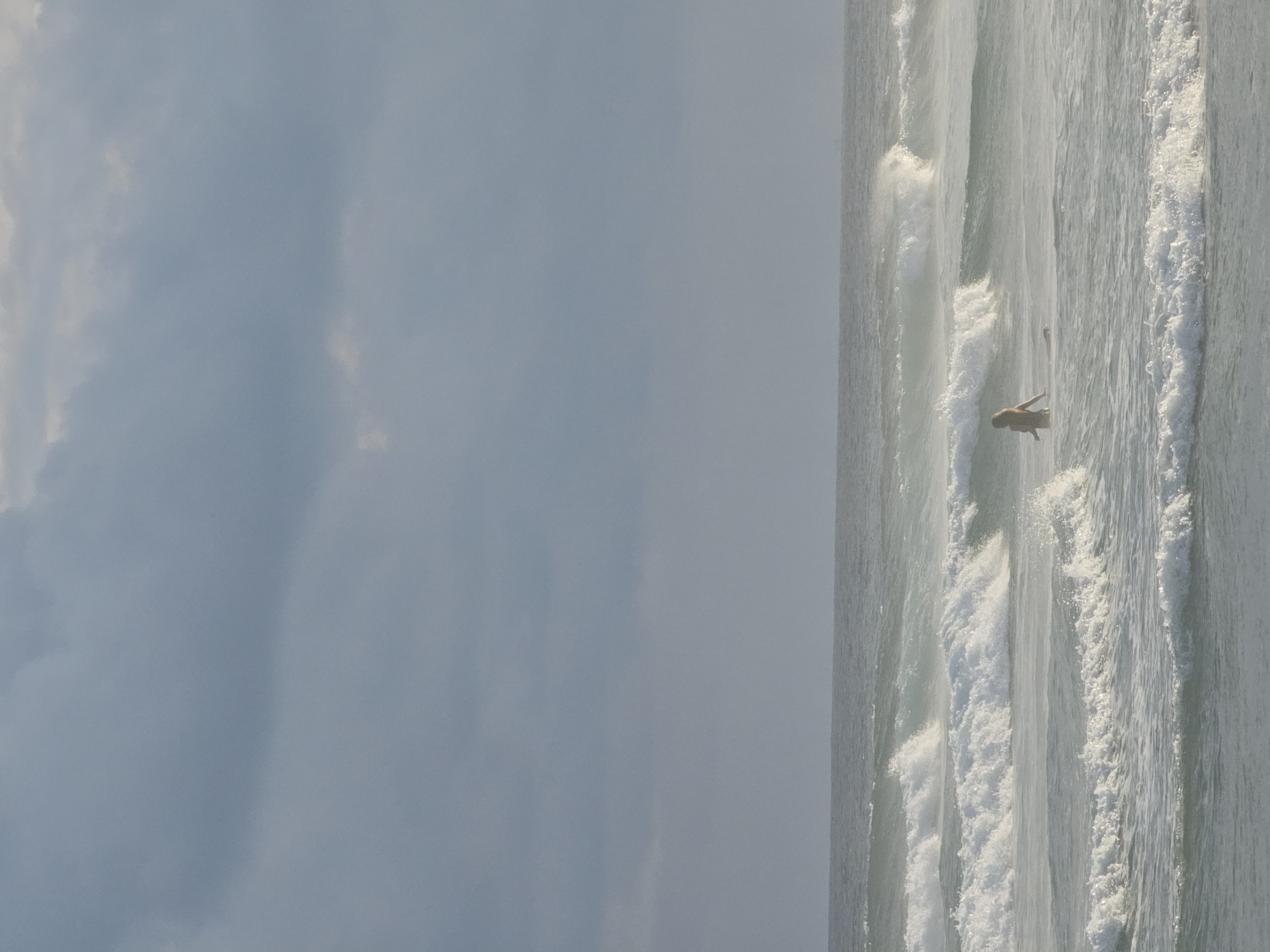 The image depicts an ocean scene under a cloudy sky. The ocean's surface is marked by multiple rows of waves, with foamy white crests. A person is seen in the water, amidst the waves. The image suggests a coastal environment. There is no information to confirm this image is from Kathaluwa, Sri Lanka.