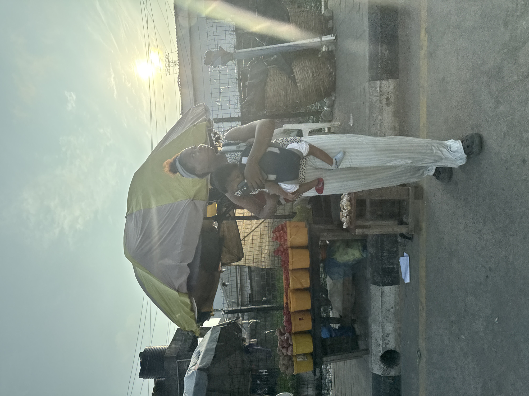 The image depicts a woman carrying a baby in a carrier as she walks past a market stall. The woman has her hair tied up and is wearing a patterned top and striped pants. The baby is wearing a white onesie and blue socks. The market stall is set up outdoors under a striped umbrella and displays various produce in yellow buckets, including what appears to be yams, tomatoes, and possibly other vegetables. In the background, there are woven baskets, a fence, and some utility poles with wires. The scene appears to be in a somewhat dusty or unpaved area, with a concrete gutter and asphalt road in the foreground. The sun is visible in the sky, suggesting it is daytime, possibly late afternoon given the angle of the light. The location is identified as Igboefon, Nigeria.