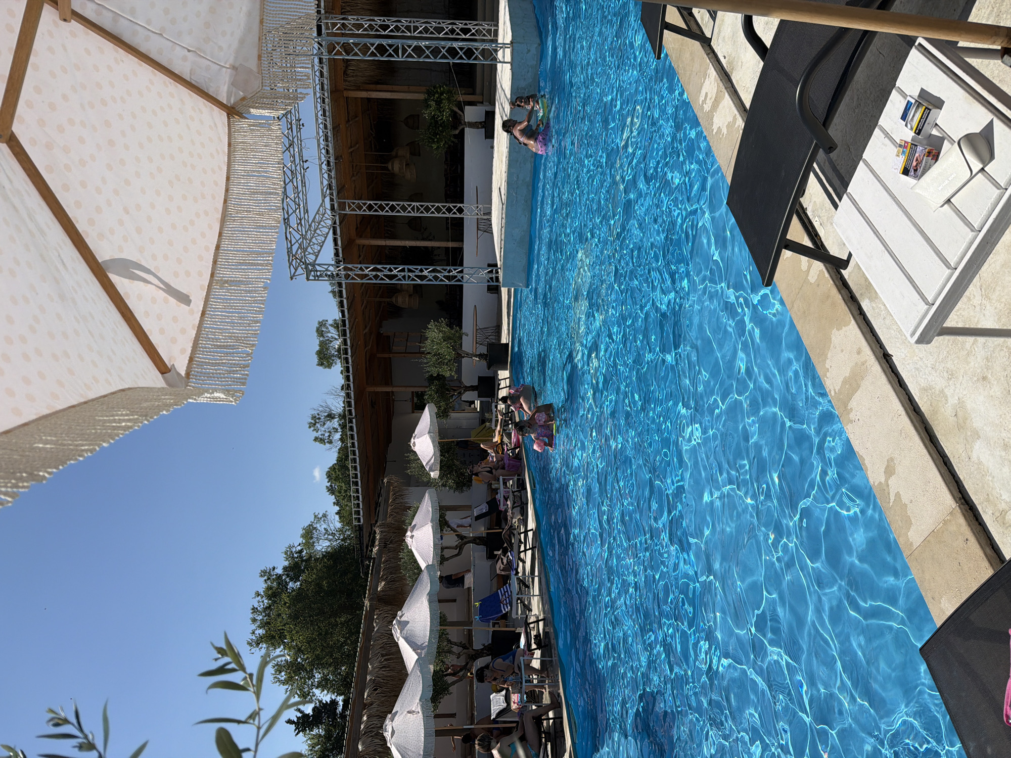 The image depicts a sun-drenched outdoor swimming pool area, likely at a resort or a beach club, on a clear, bright day. The pool itself is a vibrant, deep blue, with sunlight creating shimmering patterns on its surface. Several lounge chairs are positioned around the pool, some occupied by people relaxing. There are also large, decorative umbrellas providing shade. In the background, a covered structure with a thatched roof and exposed wooden beams suggests a bar or restaurant area. The scene appears to be in a warm climate, given the foliage and attire of the people. The specific location of Burgas, Bulgaria is not discernible from the visual cues in the image; no landmarks or signage indicate this city. The image captures a leisurely atmosphere, with people enjoying the water and the sunny weather.