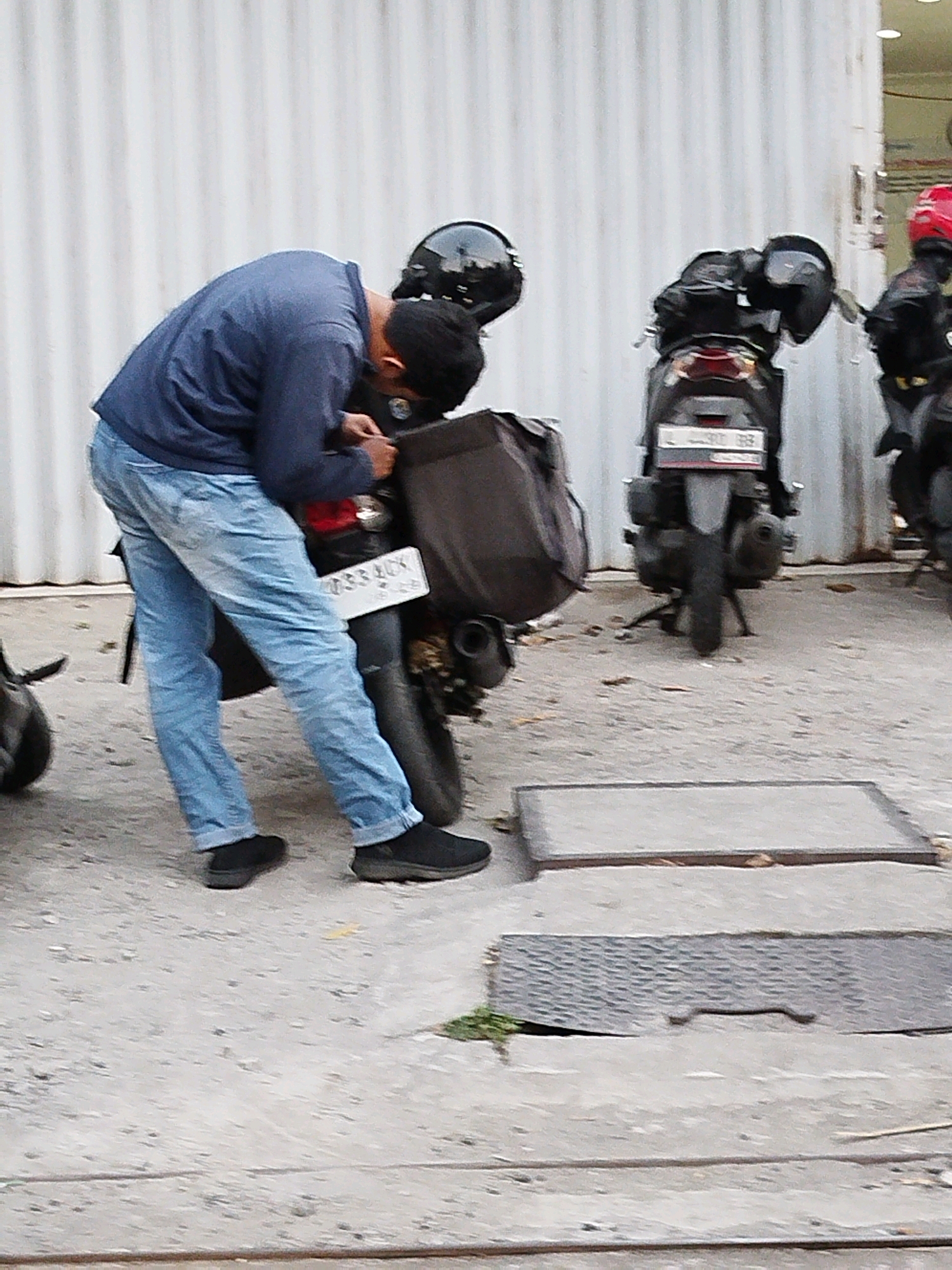 The image depicts a street scene in Surabaya, Indonesia, with a focus on two motorcycles and a person interacting with one of them. The background features a white corrugated metal wall.
In the foreground, a person wearing a dark blue long-sleeved shirt and light blue jeans is bent over a dark-colored motorcycle. The person's head is lowered, obscuring their face, and their hands appear to be near the motorcycle's seat or storage compartment. Black slip-on shoes are visible on their feet. The motorcycle has a dark storage bag attached to its rear. A license plate is visible on the back of this motorcycle, displaying alphanumeric characters that are partially obscured and difficult to read. Another dark motorcycle is parked next to it, with a black helmet resting on its seat.
Further to the right, a third motorcycle is partially visible. This motorcycle is black and has a rider wearing a red helmet and a black jacket. The license plate on this motorcycle is also difficult to discern.
The ground is paved with concrete, and two rectangular metal grates are present. Sparse dry leaves are scattered on the ground. The lighting suggests an outdoor setting, possibly daytime. The overall impression is a snapshot of everyday urban life in Surabaya.