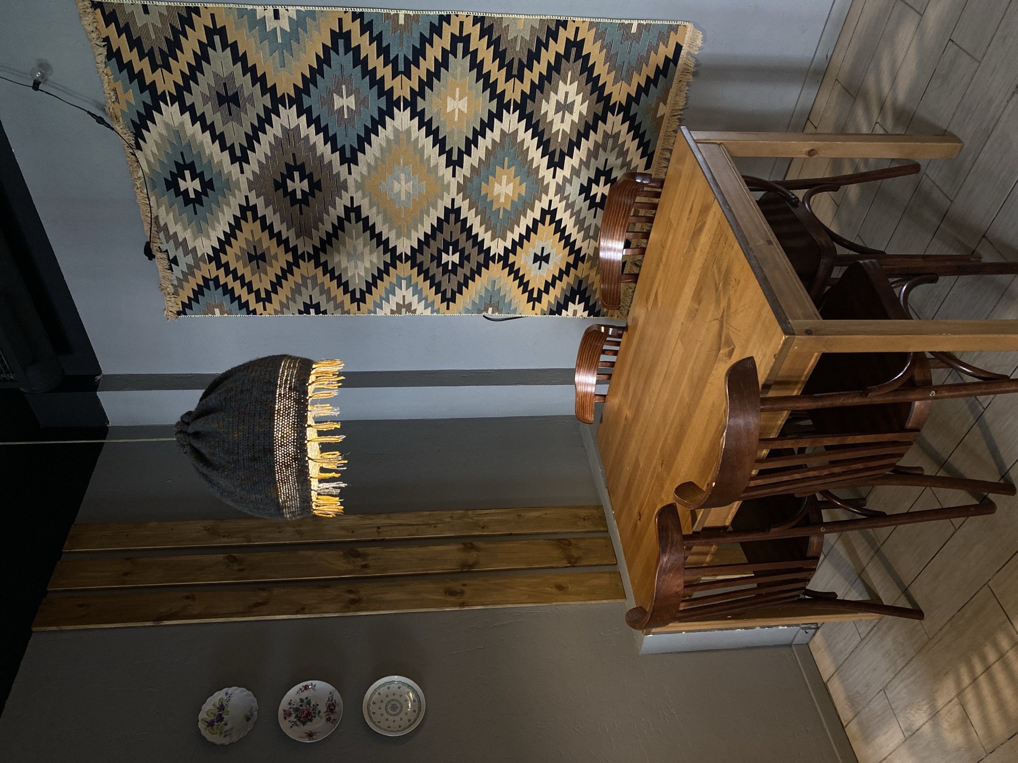 This image captures an interior scene, likely a dining area or a cafe, presented from a tilted perspective. The most prominent feature is a large, geometrically patterned rug affixed to the ceiling. This rug displays a kilim-style design with a repeating pattern of diamonds and zigzag lines in shades of blue, gold, black, and cream, with fringed edges.
A rectangular wooden dining table with a light-colored top occupies the right side of the frame. Several dark wooden chairs, consistent with a Bentwood style, are arranged around the table. Some chairs are partially stacked or pushed together.
In the mid-ground, a distinctive pendant lamp hangs. It features a woven or knitted grey-textured shade, from which warm, bright light emanates, highlighting a fringe-like detail at its base.
The walls are painted a plain medium grey or greige. On the lower left portion of the wall, three decorative ceramic plates with delicate floral patterns are hung. The floor, visible towards the bottom right, is covered with light-toned tiles, possibly designed to resemble wood planking.
There are no people or any specific activity depicted in the still scene. The lit lamp suggests the photograph was taken indoors during the evening or in a space requiring artificial illumination. The overall ambiance combines rustic and traditional decorative elements. No visible text appears in the image. The setting is located in Saint Petersburg, Russia.