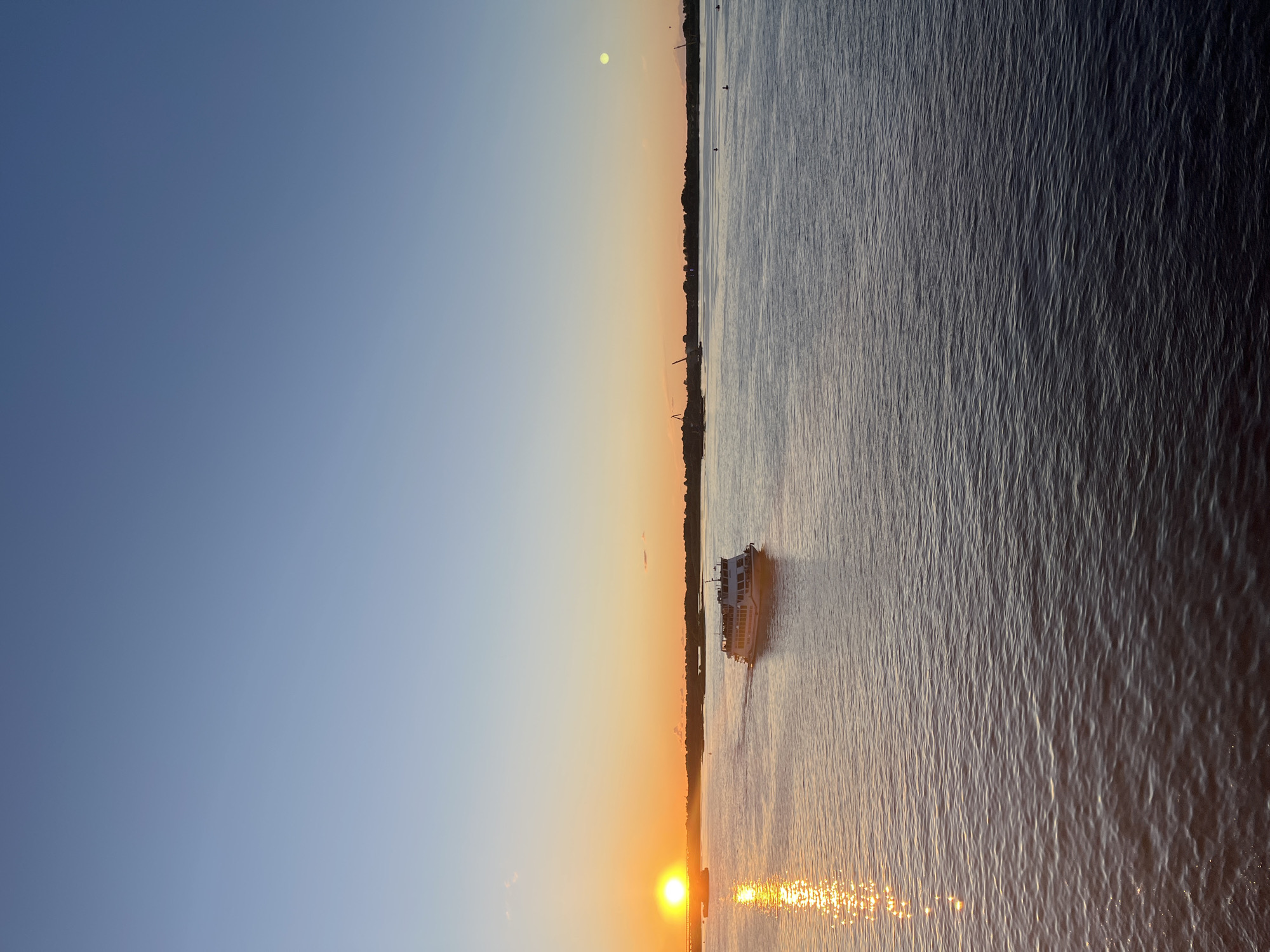 The image depicts a serene sunset over a wide body of water, likely the Volga River, in Nizhny Novgorod, Russia. The sun, a brilliant orb, is low on the horizon, casting a warm golden glow that reflects off the rippling water. The sky transitions from a deep blue at the top to a soft orange hue near the horizon, with a few wispy clouds scattered across the expanse.
A white ferryboat is the most prominent subject in the middle of the river, its wake trailing behind as it moves away from the viewer. The ferry appears to be carrying passengers, though they are not clearly discernible. In the distance, the silhouette of the city's shoreline is visible, marked by trees and the skeletal structures of cranes, suggesting a port or industrial area. The water itself is calm, with gentle waves reflecting the fading light. The overall impression is one of tranquility and natural beauty, enhanced by the soft, diffused light of dusk.