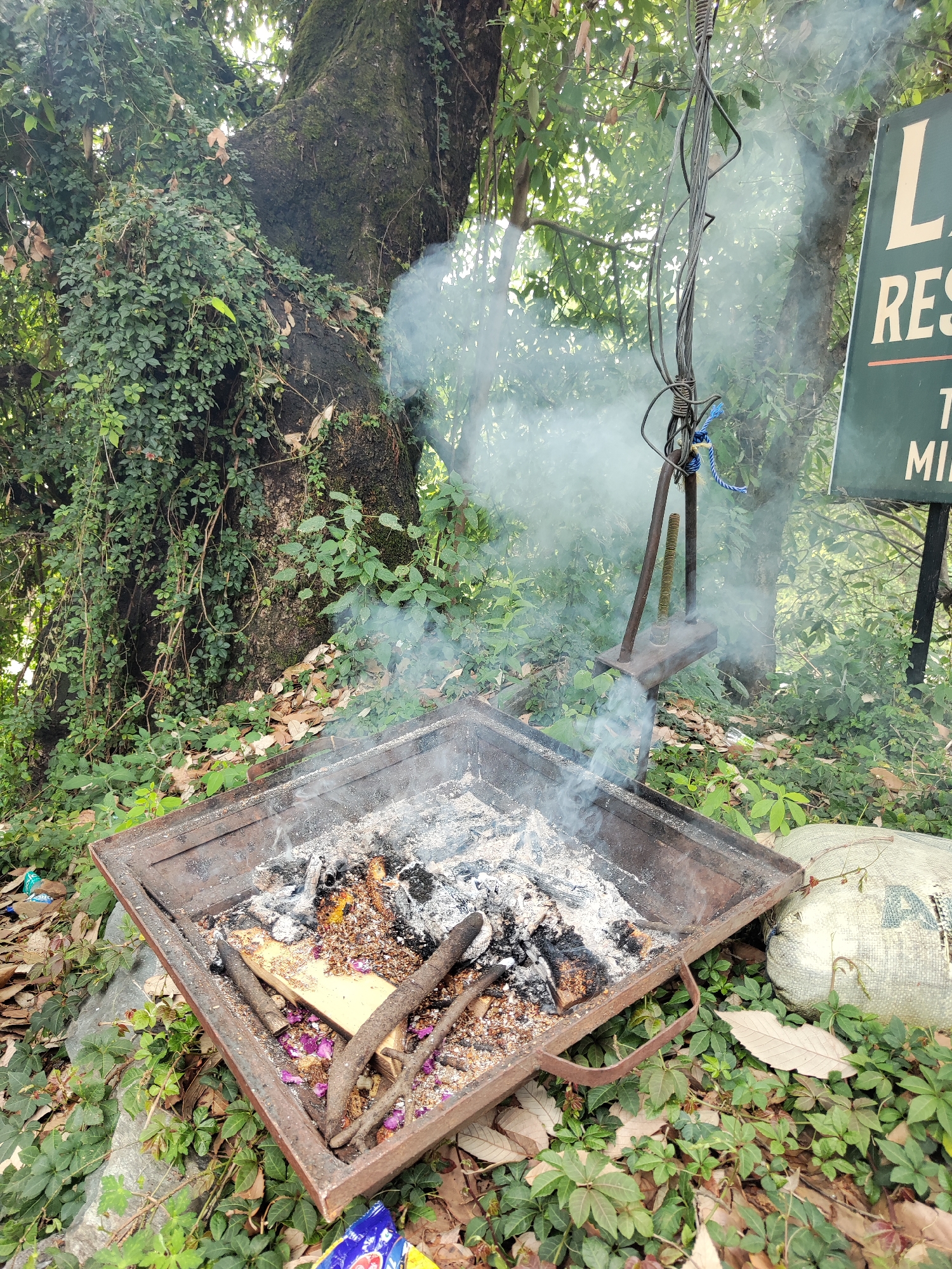 The image depicts a square metal fire pit placed on ground cover of small leafy plants in an outdoor environment. Inside the fire pit, there are burnt wood fragments and ashes. Smoke is rising from the pit. Behind the pit, there's a metal structure with cables and a metal rod, with a tree and a sign with the words "RESTAURANT" visible in the background. A white burlap bag and discarded wrappers are also visible on the ground.