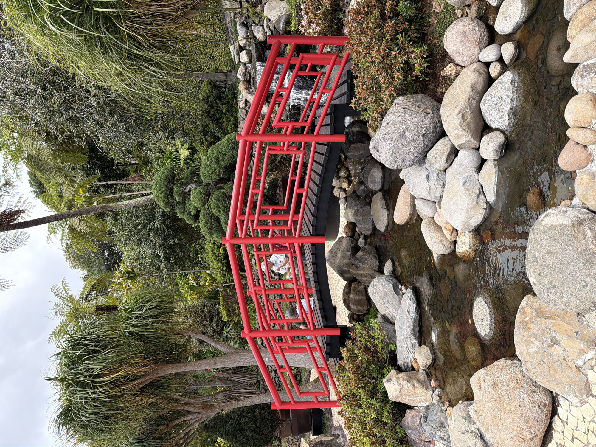 The image shows a Japanese garden located in the city of Funchal, Portugal. A red bridge crosses a stream bed of rocks. A small waterfall is visible beneath the bridge. Trees, bushes, and foliage surround the bridge and stream. Two people are on the bridge.
