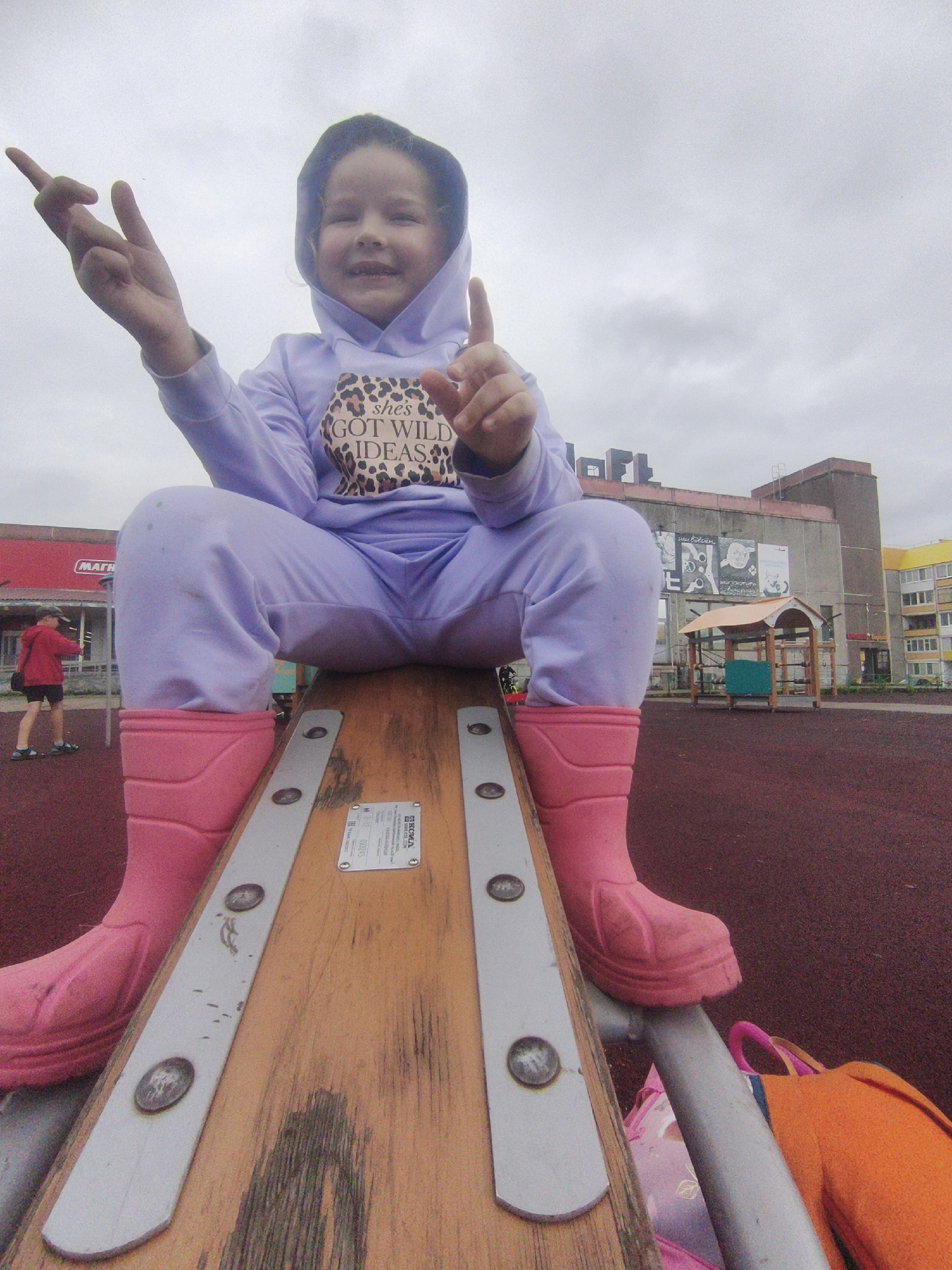 A low-angle, wide shot captures a young girl sitting on a seesaw in a playground. She is wearing a light purple hooded sweatshirt with "she's GOT WILD IDEAS" printed on it in a leopard-print font, and matching light purple sweatpants tucked into bright pink rubber boots. Her arms are raised, and she is holding up two fingers on her left hand and one finger on her right hand, as if making a gesture. Her expression is cheerful, with a wide smile.
The seesaw is made of wood with metal reinforcements. The playground surface is a deep red, possibly rubberized material. In the background, there's a building with several large posters or murals on its exterior, and further in the distance, a residential building with yellow and white balconies. A small wooden playground structure with a light brown roof and green panels is visible to the right of the building. To the left, a boy in a red jacket and black shorts walks on the playground surface.
The sky is overcast and gray, suggesting a cloudy day. The location is identified as Марковский, Russia. The overall mood is playful and bright, despite the cloudy weather. Visible text includes the writing on the girl's sweatshirt and a label on the seesaw which appears to be in Cyrillic, with the word "ТЕХНИКА" (Tekhnika - Technology/Equipment) and some numbers.