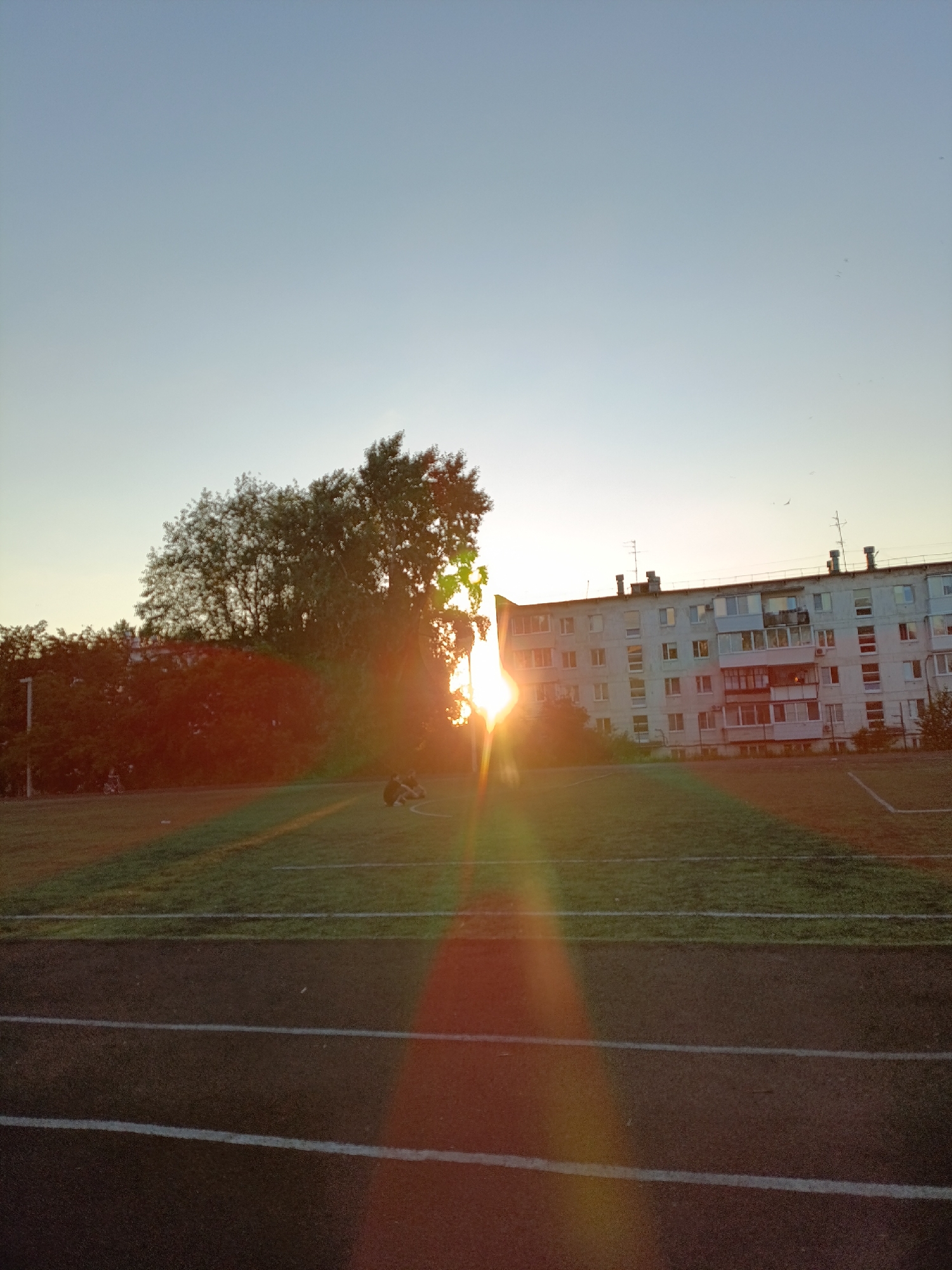 The image depicts a late afternoon or early evening scene on a grassy field, possibly a sports field, in the city of Chaikovsky, Russia. The sun is low in the sky, casting a strong lens flare that obscures part of the scene. In the background, a multi-story apartment building stands, with several antennas and chimneys visible on its roof. Trees, some dense and others more sparse, are situated between the field and the building. The foreground shows a dark running track with white lines marking lanes. Two individuals are sitting on the grass in the middle ground, facing away from the camera, with their legs stretched out. The sky is clear and a pale blue, transitioning to a lighter hue near the horizon where the sun is setting. The overall impression is one of a quiet, everyday scene in a residential area of Chaikovsky during a period of low sunlight.