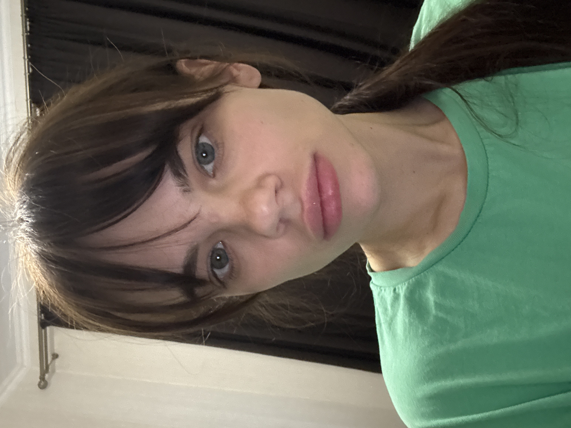 A woman with brown hair and green eyes is pictured from a low angle. She has bangs that partially cover her forehead. She is wearing a green t-shirt. The background is dark, with what appears to be a curtain or draped fabric. A white wall with a metal rod is visible in the lower left corner. The image does not depict the location of Rostov-on-Don, Russia.