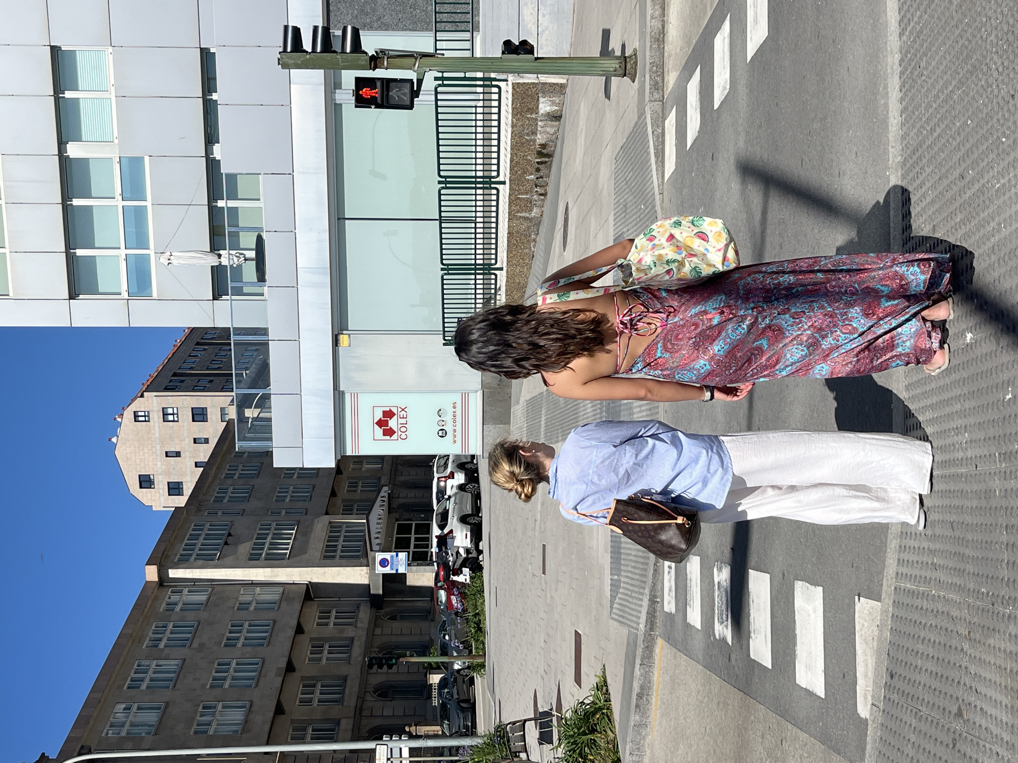 Two women are walking across a pedestrian crossing in a city, possibly A Coruña, Spain. The woman in the foreground is wearing a light blue striped shirt and white pants, carrying a brown designer handbag. She has blonde hair pulled back in a bun. The woman behind her is wearing a long, colorful patterned dress and a printed tote bag. She has dark, wavy hair. They are walking on a sunny day, as indicated by the strong shadows. The setting includes modern buildings with glass facades and older architecture, suggesting an urban environment. A traffic light showing a red pedestrian signal is visible. There is text on a sign that reads "COLEX" with a website address "www.colex.es". The scene appears to be in the daytime.