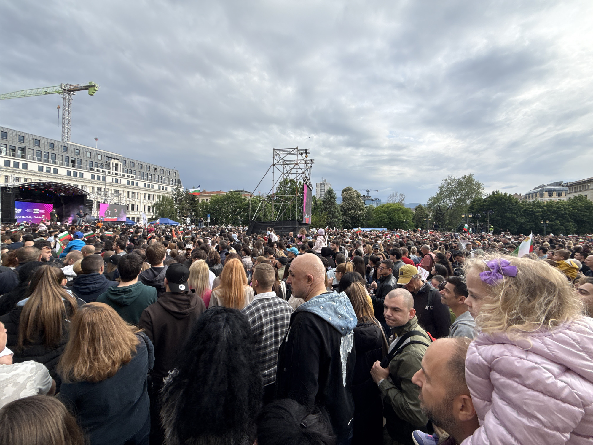A wide-angle shot shows a large crowd of people gathered in a square in Sofia, Bulgaria. The crowd extends throughout the frame. In the background is a stage with a screen and a large building with a construction crane beside it. There is a tall metal structure in the distance to the right of the stage. The sky is cloudy. In the foreground, people are of varying heights and dressed in various clothing, including a girl in a pink jacket with a purple bow in her hair being carried on someone's shoulders. There are some Bulgarian flags scattered throughout the crowd.