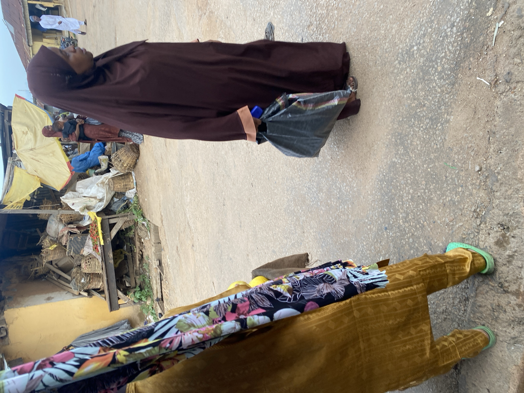 The image depicts a street scene in Jos, Nigeria. The primary subject is a woman wearing a dark brown hijab and a traditional brown garment. She is walking on a paved street, holding a black plastic bag. In the background, there are market stalls with various items, including woven baskets and yellow tarps. Another person, possibly a woman, is visible in the distance wearing a red and white patterned headscarf and a brown skirt. Further in the background, a man in a white traditional outfit is walking. The ground is a mix of concrete and dirt, with some debris scattered around. The scene suggests a bustling marketplace environment, typical of a daytime setting in Nigeria. The lighting indicates it is daytime, and the overall atmosphere appears to be that of everyday commerce and pedestrian activity. There is no visible text in the image.