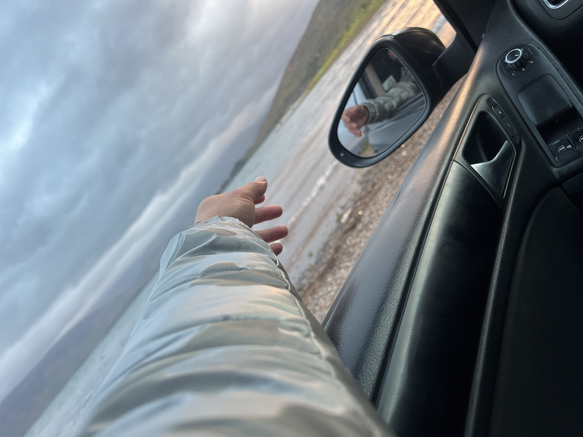 From inside a car, a person extends a hand towards a body of water under a cloudy sky. The shoreline is visible. The car's side mirror reflects an arm. The car's interior includes a door panel with controls. The location is not discernible from the provided image to determine the city of Bin El Ouidane, Morocco.