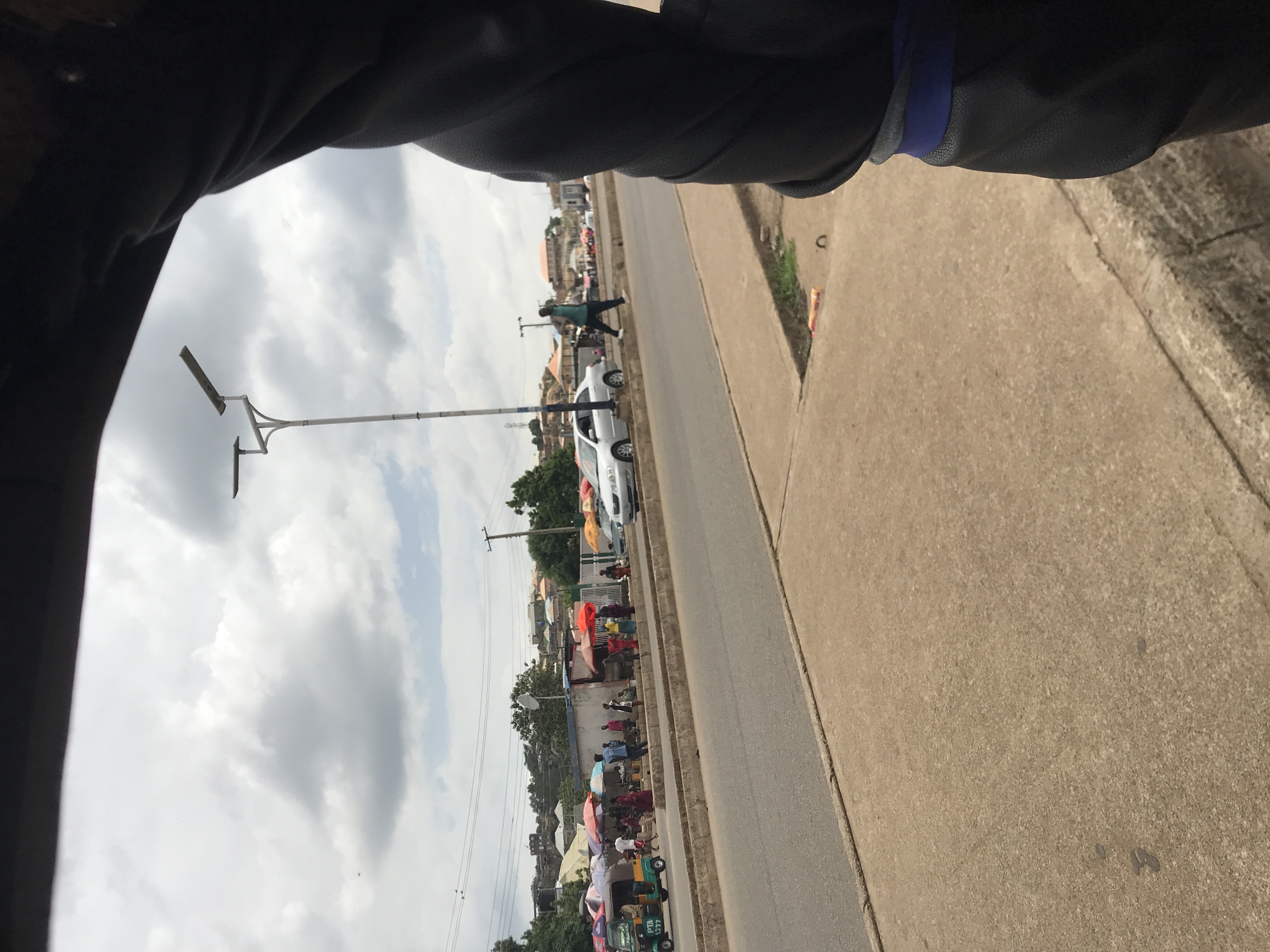 The image captures an outdoor street scene in Kabong, Nigeria, under an overcast sky filled with grey and white clouds. The perspective is slightly angled, looking across a multi-lane asphalt road towards a commercial or market area on the left.
In the foreground and to the right, a wide, light-brown concrete sidewalk or shoulder is visible, with a dark, textured object (possibly a vehicle interior or arm) occupying the far right edge of the frame, showing a blue and grey strap. A concrete curb separates the sidewalk from the road.
The asphalt road features a light-colored concrete median separating opposing traffic lanes. On the far side of the road, another concrete strip borders a pedestrian area. A tall, white street light pole with two horizontal, rectangular light fixtures stands in the median towards the center-left.
Numerous individuals are present on the left side of the road, congregating around stalls shaded by colorful market umbrellas (red, blue, orange, green). A green and yellow trishaw, marked "PTA 1777," is visible on the far left of the road, near the market activity. Further down the left lane, a silver sedan is parked or moving slowly. Another silver car is partially visible behind it.
One man, wearing a green shirt and dark trousers, is captured in motion, striding across the road from left to right. In the background, beyond the market, a low, light-colored building is visible, with utility poles and overhead wires above it. Distant hills or elevated terrain are dotted with numerous residential structures under the cloudy sky.