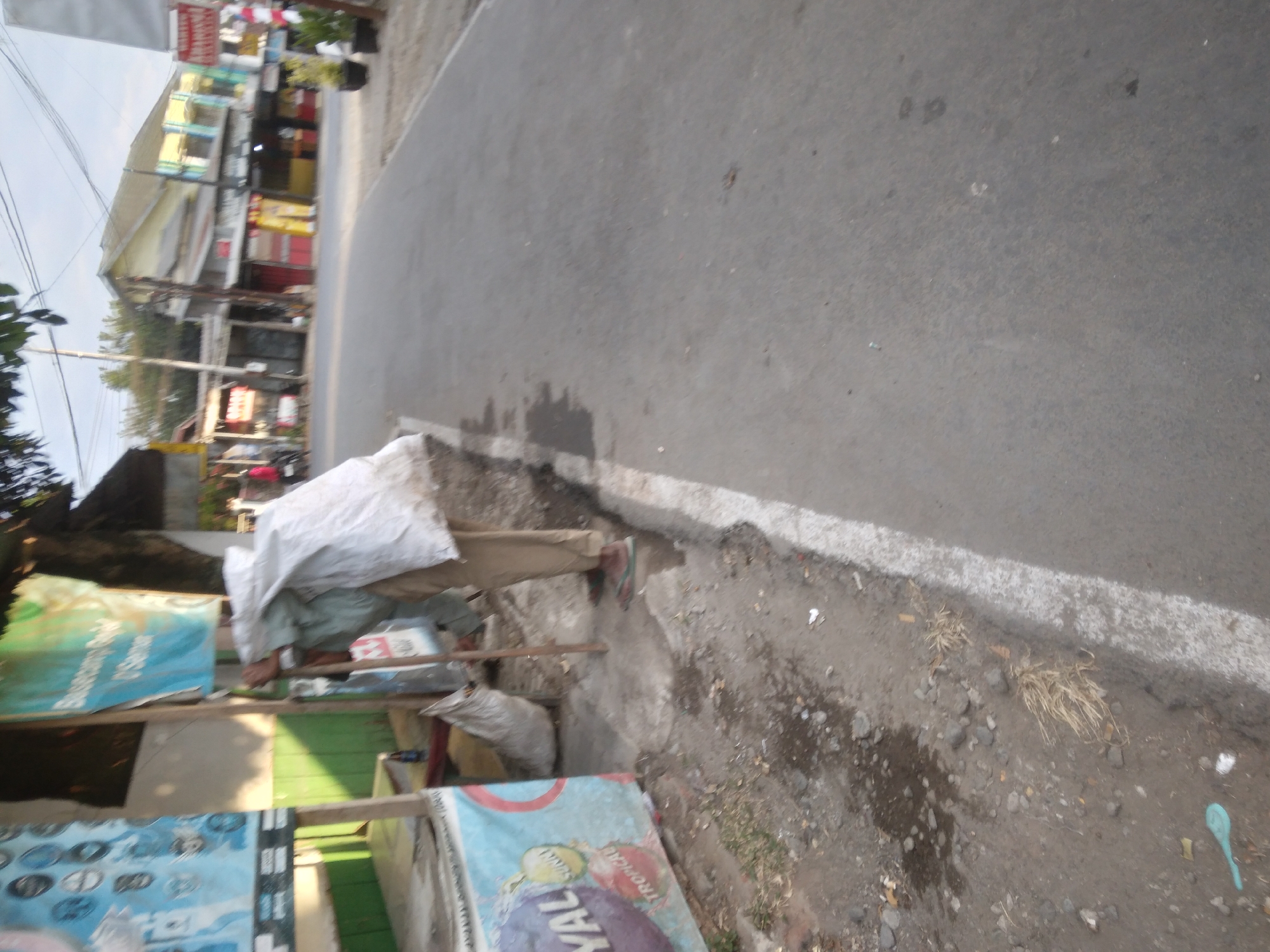 A person wearing a light green shirt and beige pants is standing on a street in Desa Pamijen, Kecamatan Sokaraja, Indonesia, carrying a large white sack over their shoulder. The person is holding a long stick or pole with their right hand, which is also supporting the sack. A dirty, greyish-white sack is lying on the ground to the left of the person. The area appears to be a roadside, with a paved road visible on the right and a rough, uneven ground with some debris on the left. There is a blue and white patterned cooler or display unit to the far left, with some text and images on it. Further back, a row of buildings with various storefronts lines the opposite side of the street, some with yellow and red awnings. Trees and foliage are visible behind the buildings. The sky is overcast.