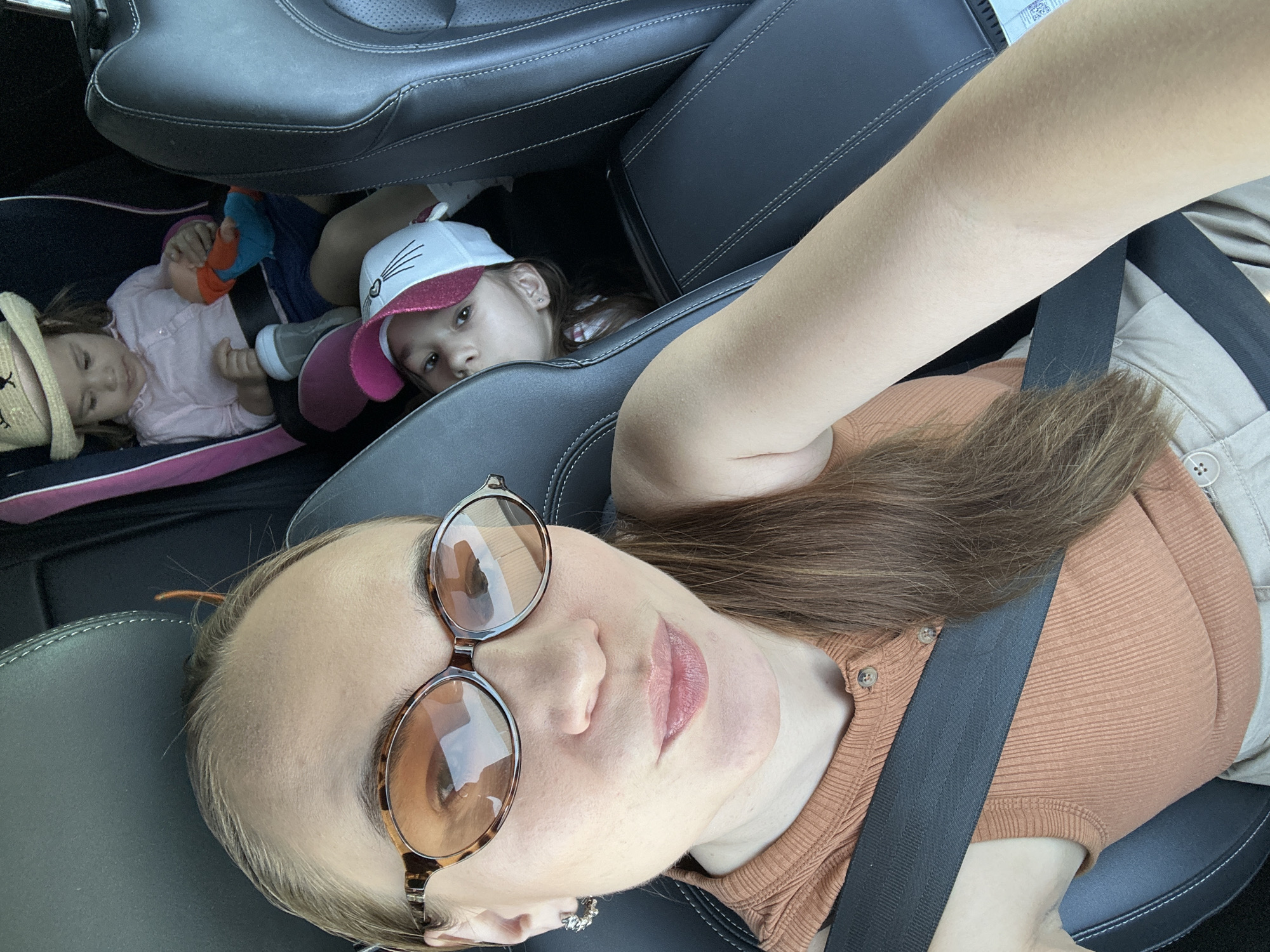 A selfie-style, slightly tilted, medium shot shows a woman in a car with two children in the back seat. The woman is wearing a brown ribbed shirt, beige pants, and a seatbelt. She has light skin, brown hair, and sunglasses resting on her forehead. The background behind her contains two young children in a car seat, who appear to be sleeping or resting. The location of this image is unknown and there is no verifiable evidence to suggest that it was taken in the city of Kharnavy, Russia.