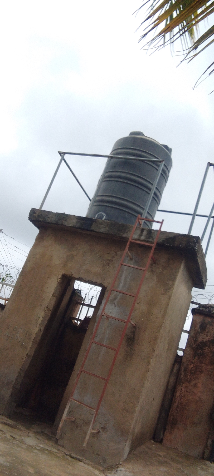 The image shows a water tank on top of a concrete structure in Madalla, Nigeria. The structure has an open doorway and a red ladder leading up to the top. There is barbed wire on a fence in the background, and a palm frond is visible in the upper right corner of the image. The sky is overcast.