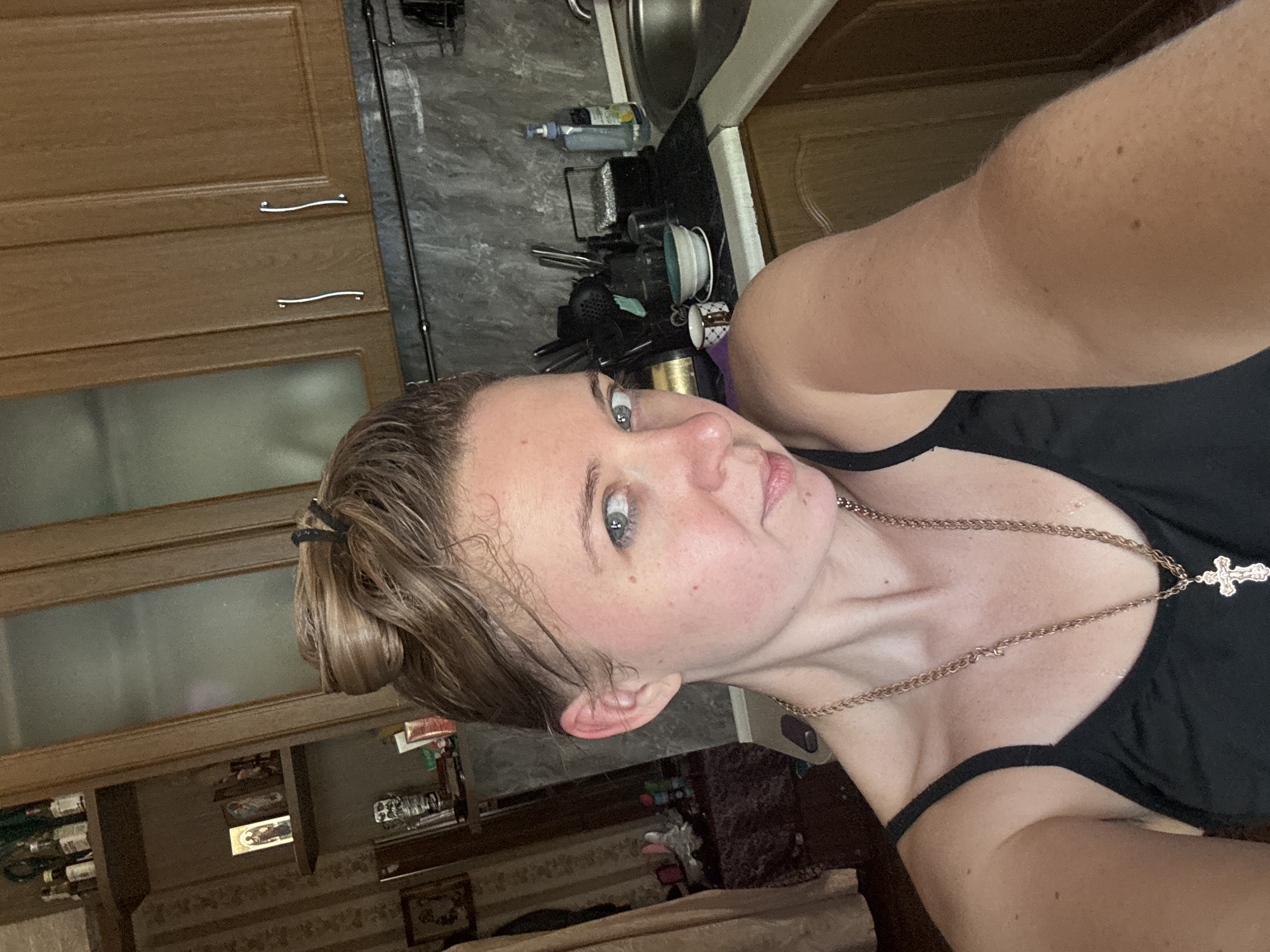 A selfie taken from a low angle shows a woman with blonde hair pulled up in a bun and blue eyes looking at the camera. She is wearing a black tank top and a gold necklace with a cross pendant. The background appears to be a kitchen with wooden cabinets and a sink. The lighting is dim, suggesting the photo was taken indoors.